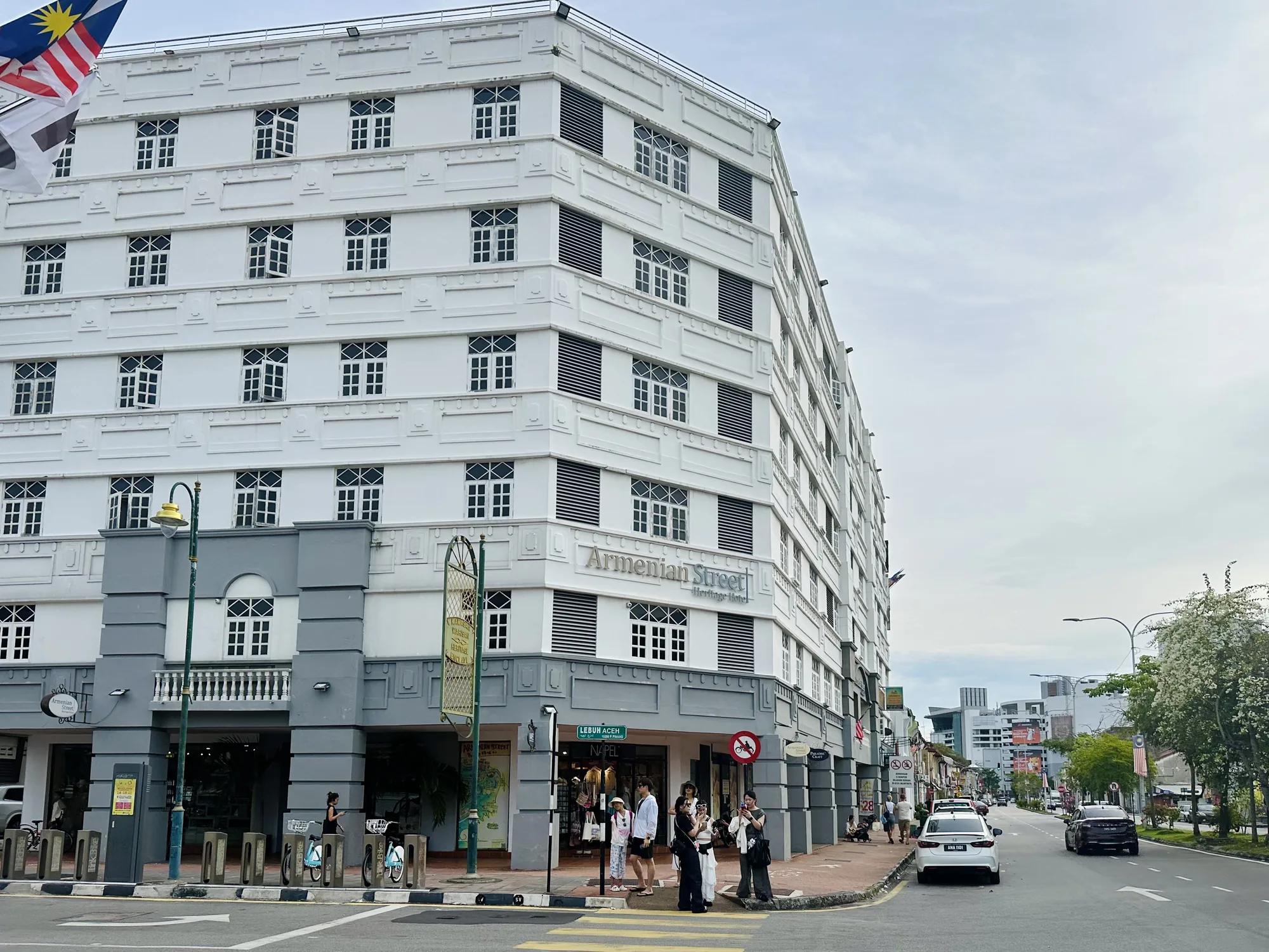 マレーシアのペナンで撮影された写真です。画面左手には白とグレーで統一された、装飾的な建物が写っています。建物には「Armenian Street Heritage Hotel」と書かれており、ホテルのようです。建物の前には歩道があり、数人が立っています。横断歩道の手前で立ち止まっている人もいれば、通りを歩いている人もいます。自転車のレンタルステーションのようなものも見えます。  画面右奥には、街路樹が植えられた道路が奥へと続いています。道路には数台の車が走行、または停車しています。空は曇りがちで、全体的に落ち着いた雰囲気の写真です。建物のデザインや看板などから、ペナンの歴史的な街並みを捉えた一枚であることがわかります。