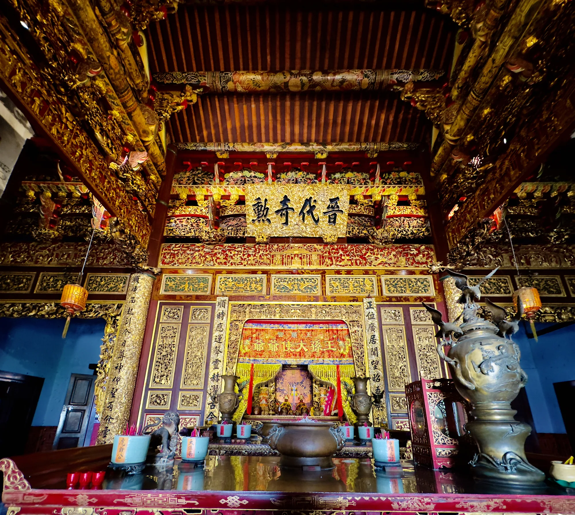 これはマレーシアのペナンで撮影された寺院の内部の写真です。写真の中心には、豪華な装飾が施された祭壇があります。祭壇は、金色の彫刻で覆われており、非常に精巧な作りです。祭壇の上には、漢字が書かれた額が掲げられています。額の文字は「二動奇代晋」と読めます。祭壇には、線香立てや花瓶、その他のお供え物が置かれています。祭壇の前には、大きな香炉が置かれており、香炉の周りにも線香が立てられています。天井は木製で、赤茶色に塗られており、細かく装飾されています。全体的に、寺院は厳かで、豪華な雰囲気を醸し出しています。写真から、この寺院が地域の文化や信仰の中心地であることが伝わってきます。祭壇の左側には青色の壁が見え、右側には青銅の鷲の像が置かれているのが確認できます。