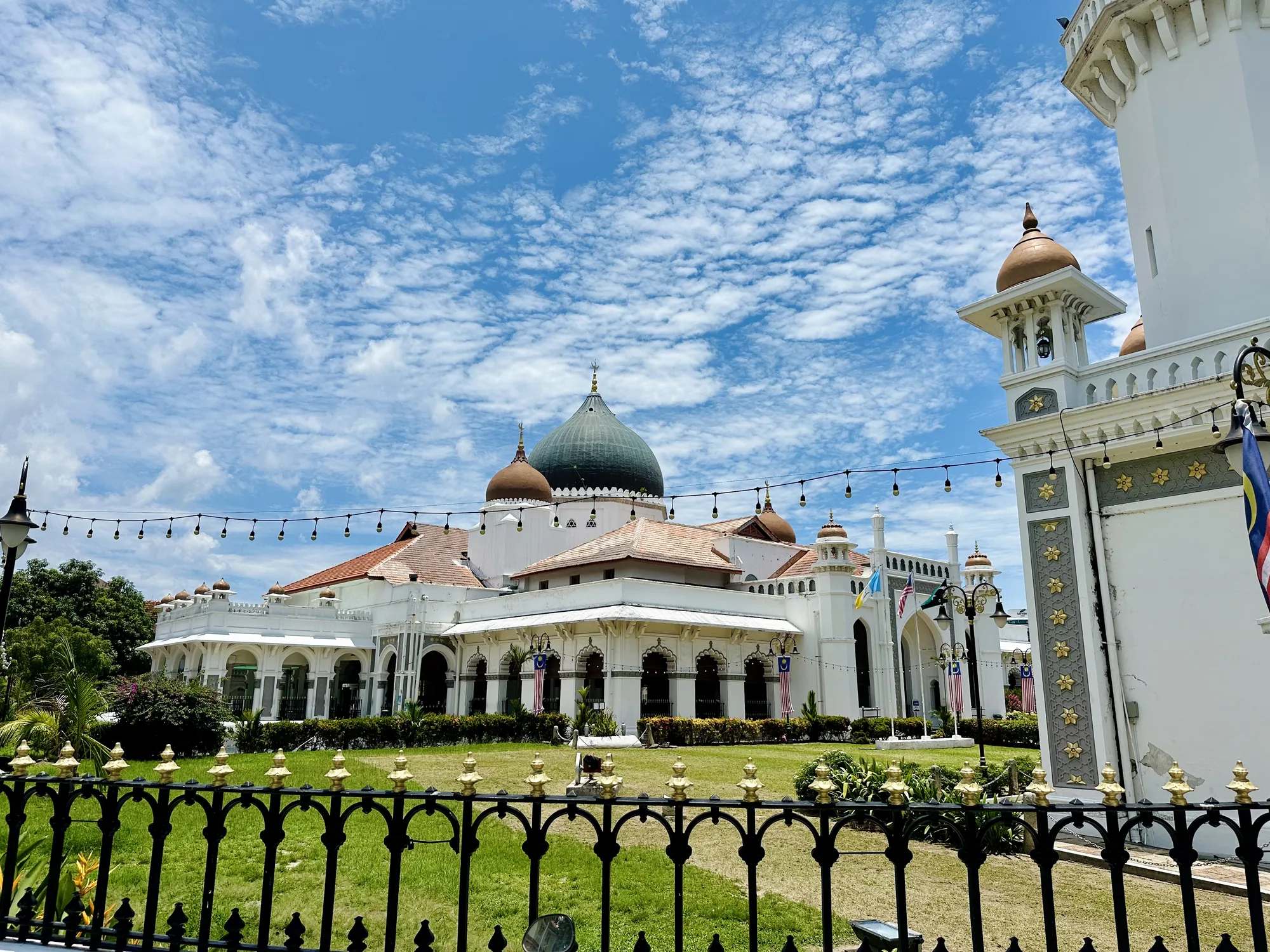写真はマレーシア、ペナンにあるモスクを捉えたものです。空には白い雲が広がり、強い日差しが照りつけています。前景には黒色の柵があり、金色の装飾が施されています。柵の向こうには緑の芝生が広がり、丁寧に手入れされている様子が伺えます。中央には白を基調とした壮麗なモスクがそびえ立ち、建物の屋根は赤茶色、ドームは緑色をしています。モスクの壁面にはアーチ状の窓が並び、マレーシアの国旗が掲げられています。建物の周りには街灯が設置され、電球が連なっています。全体的に明るく、穏やかな雰囲気の写真です。