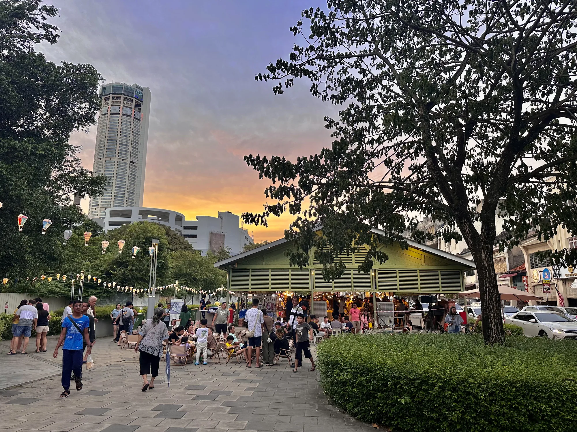 マレーシア、ペナンの夕暮れ時の写真です。空は夕焼けでオレンジ色と紫色のグラデーションになっており、美しいです。写真の中央には、人々が集まっている場所があります。小さな屋台のようなものがあり、そこで人々が飲食を楽しんでいるようです。手前には石畳の道があり、人々が歩いています。奥には高いビルが見え、その近くには木々が生い茂っています。空には提灯のようなものが吊るされており、お祭りか何かのイベントが行われているのかもしれません。全体的に穏やかで活気のある雰囲気の写真です。屋台には「豆菜 芋泥豆腐花」と書かれており、豆腐を使ったデザートを売っているようです。