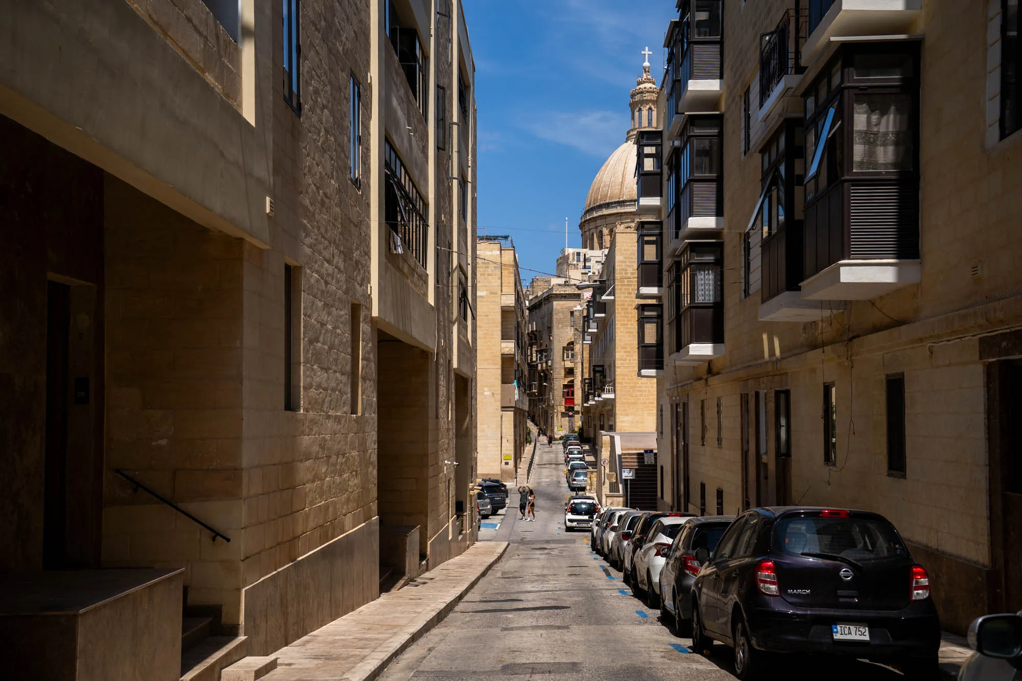 マルタで撮影された写真です。狭い路地の奥には、ドーム型の屋根を持つ教会が見えます。路地の両側には、マルタ特有の石造りの建物が並んでいます。建物はベージュ色で統一され、窓枠やバルコニーがアクセントになっています。路地の片側には、車が縦列駐車しています。路地の奥の方には、2人の人物が歩いているのが見えます。空は晴れており、建物に影が落ちています。全体的に落ち着いた雰囲気で、マルタの街並みの日常風景を切り取った写真です。