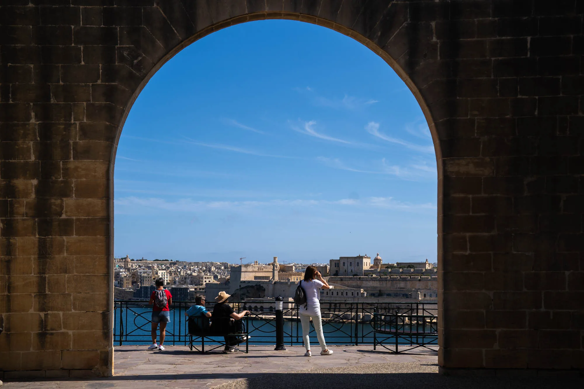 写真は、マルタの石造りのアーチから景色を眺める人々を捉えています。アーチはレンガでできており、暗い影の中にあります。アーチの向こうには、青い空と白い雲が広がり、その下には青い海が広がっています。海の向こうには、マルタの街並みが広がっており、多くの建物が見えます。手前には、ベンチに座って景色を眺める人々や、景色を撮影する女性がいます。全体的に、写真からは穏やかでリラックスした雰囲気が感じられます。