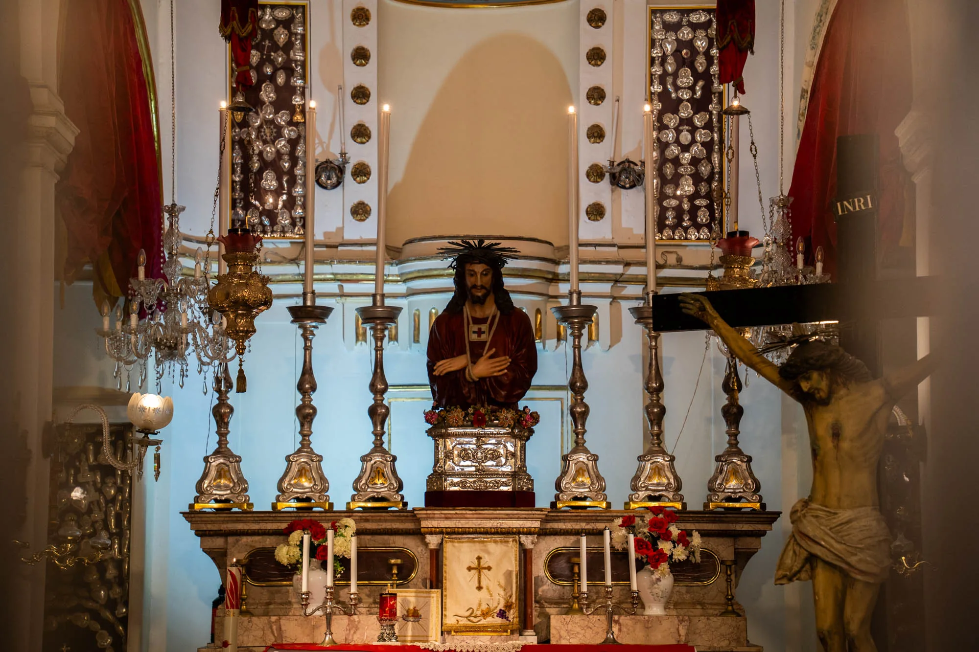 写真は、マルタの不明な場所で撮影された、教会の祭壇のクローズアップです。祭壇の中央には、茨の冠をかぶり、赤い服を着たイエス・キリストの胸像が置かれています。胸像の下には、花で飾られた銀色の台座があります。胸像の両側には、銀色の燭台が立てられ、白いロウソクが灯されています。  祭壇の下には、赤い布が敷かれており、花瓶に生けられた花や、十字架が刺繍された白い布が飾られています。祭壇の左側には、シャンデリアが吊り下げられ、赤いカーテンがかけられています。右側には、十字架にかけられたイエス・キリスト像が見えます。像の横には「INRI」と書かれた札が立てられています。  祭壇の背景には、白い壁があり、装飾的な模様が描かれています。全体的に、写真は敬虔で荘厳な雰囲気を醸し出しています。