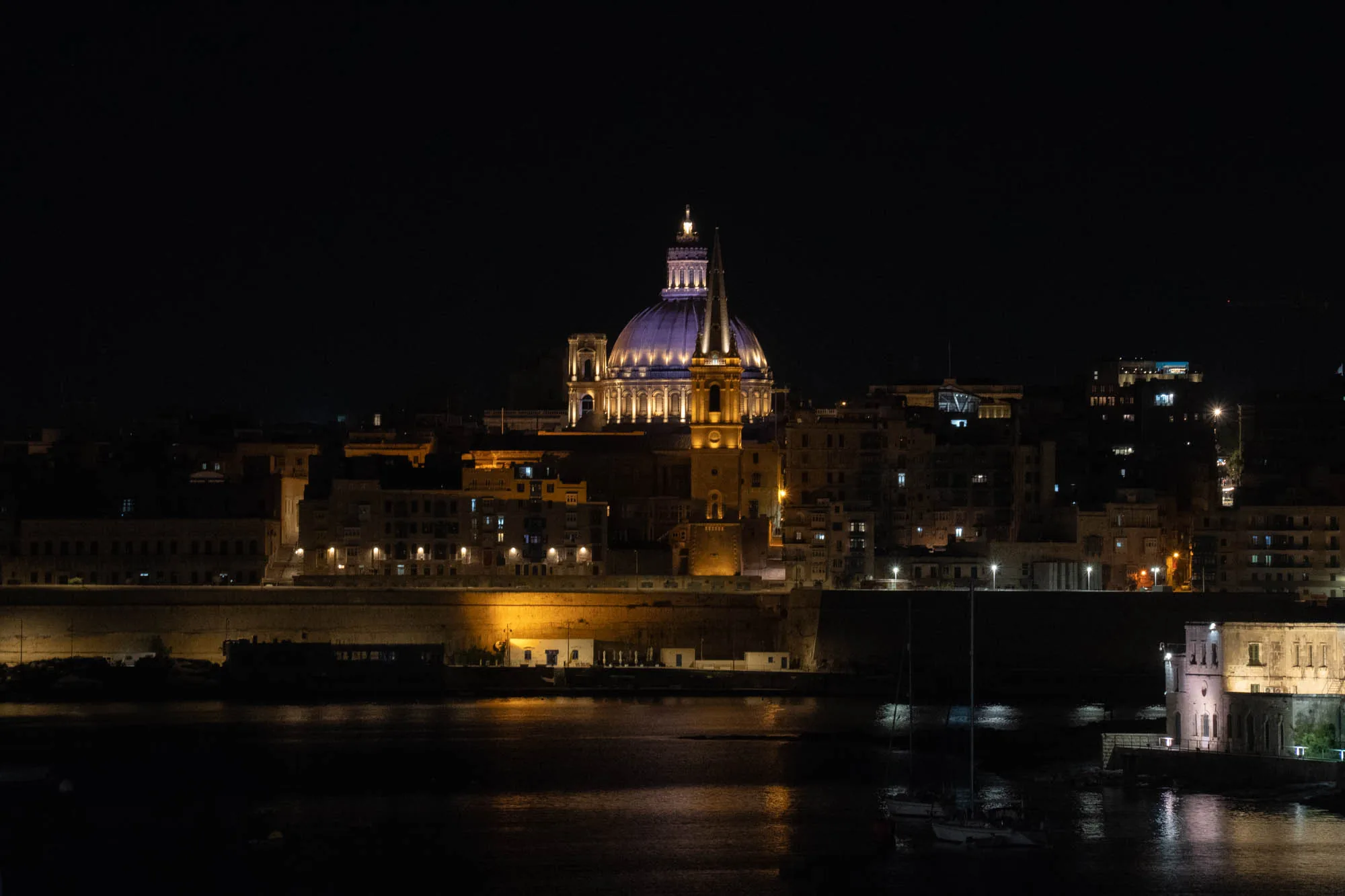 マルタの夜景を捉えた写真です。暗闇の中に、ライトアップされた聖堂が荘厳な雰囲気を醸し出しています。特にドーム型の屋根が印象的で、青みがかった光を放ち、遠くからでも存在感を示しています。聖堂の周囲には建物が立ち並び、それぞれの窓からもれる光が、街に温かみを与えています。手前には海が広がり、水面に建物の光が反射して、幻想的な光景を作り出しています。数隻のヨットが停泊している様子も見えます。全体的に落ち着いたトーンで、静かで美しい夜のマルタの風景が表現されています。