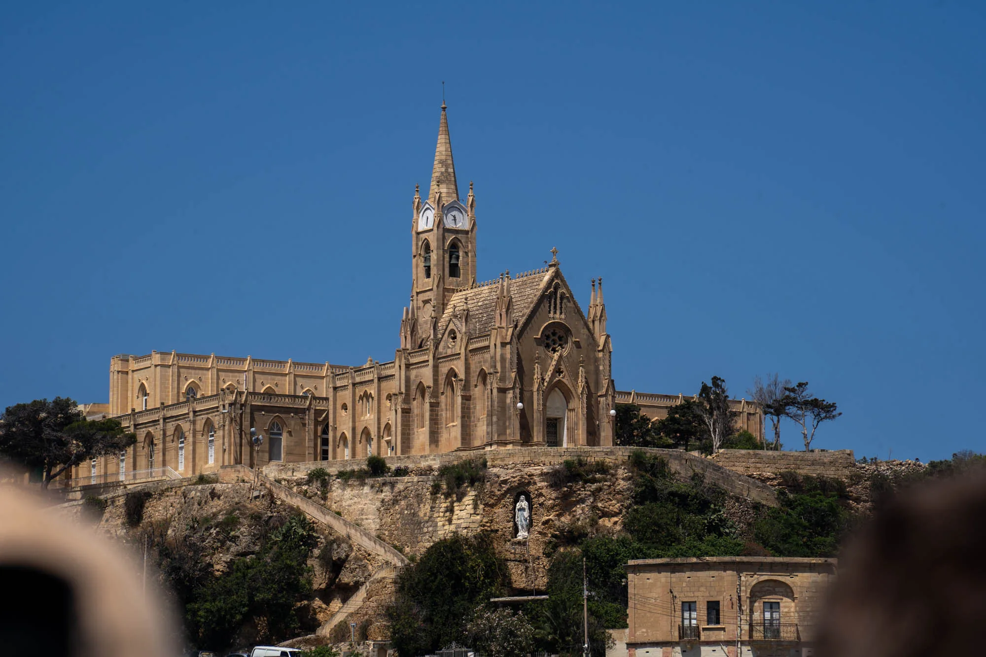 写真には、マルタにあると思われる、丘の上にそびえ立つ壮麗な教会建築が写っています。建物全体は淡いベージュ色の石造りで、ゴシック様式の特徴である尖塔や装飾が目を引きます。塔には時計が取り付けられており、建物の歴史と風格を感じさせます。教会は丘の上に建てられており、石段を登ってアクセスできるようになっています。丘の中腹には聖母マリア像が安置されているのが見えます。空は澄み切った青色で、建物のシルエットを際立たせています。手前には一部が影になっており、奥行きのある構図となっています。全体的に、マルタの美しい風景と歴史的な建造物を捉えた写真であり、静かで厳かな雰囲気が漂っています。