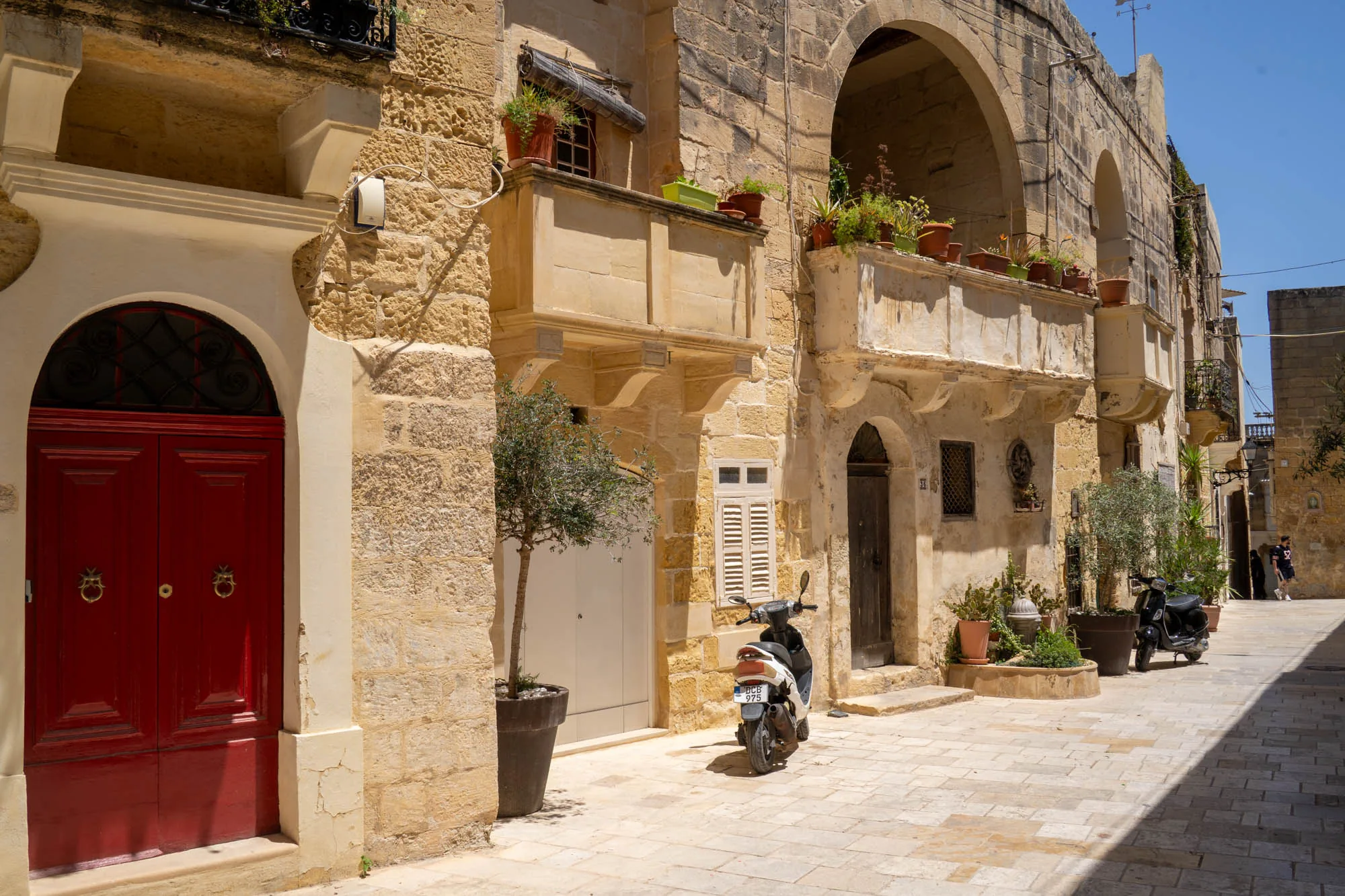 写真はマルタのどこかの街並みを捉えたものです。道の両側には、石造りの建物が並んでいます。建物はベージュ色で、窓やドア、バルコニーなどが特徴的です。一部のバルコニーには植木鉢が置かれ、緑が添えられています。道の舗装は石畳で、太陽の光が当たって陰影ができています。写真の中央にはスクーターが2台停まっています。赤色のドアやアーチ状の構造など、地中海らしい風景が広がっています。空は青く、全体的に明るく穏やかな雰囲気です。