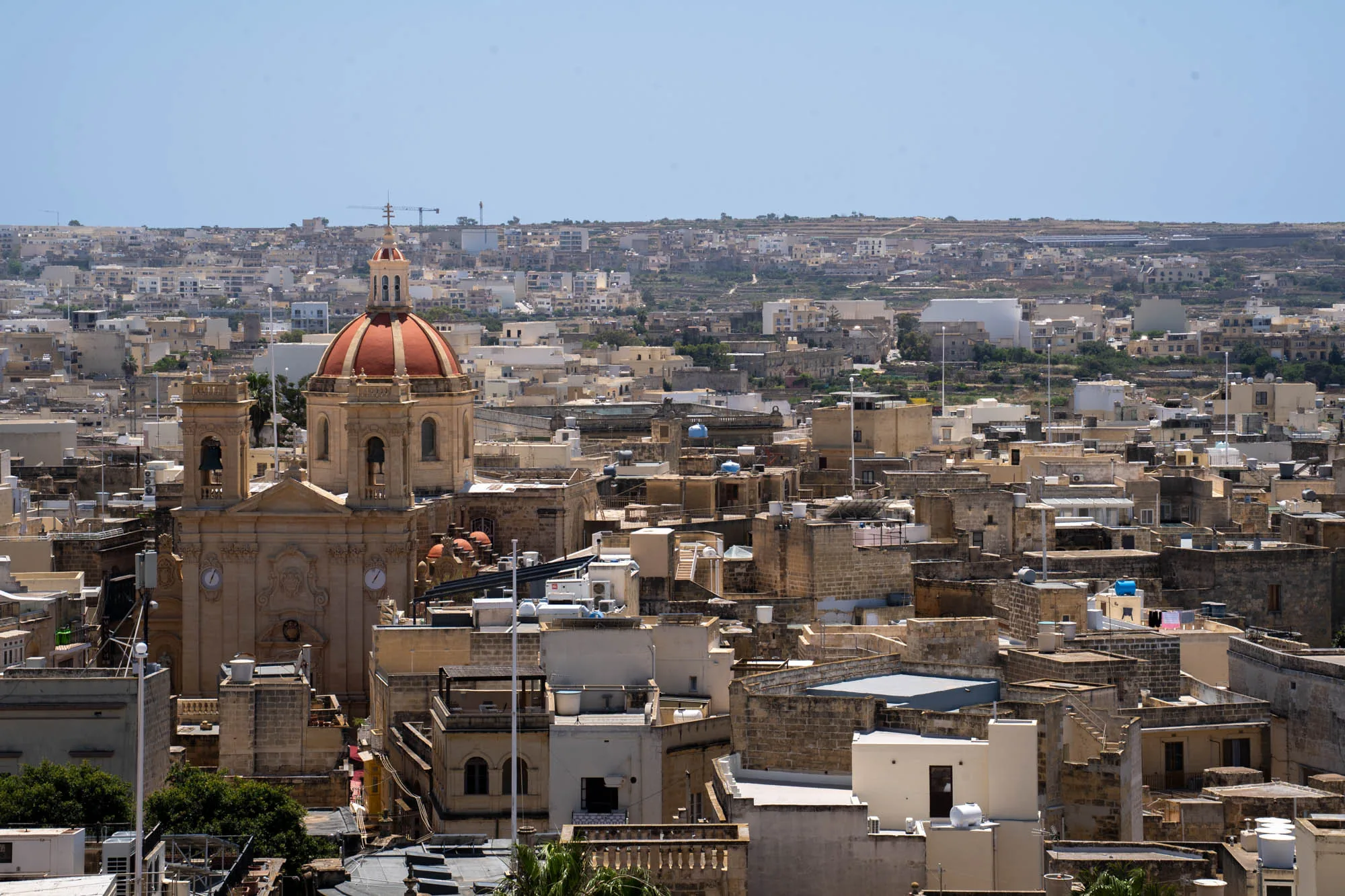 写真は、マルタの都市風景を捉えています。画面中央には、オレンジ色のドームを持つ教会がそびえ立っており、その周りをベージュ色の建物が密集して取り囲んでいます。建物は石造りで、屋上には太陽光パネルや給水タンクなどが設置されています。遠景には、さらに広大な街並みが広がり、地平線にはうっすらと丘陵地帯が見えます。空は晴れ渡っており、全体的に明るい印象です。光の加減から、昼間の時間帯に撮影されたものと思われます。建物が密集している様子から、歴史的な都市の中心部であると考えられます。