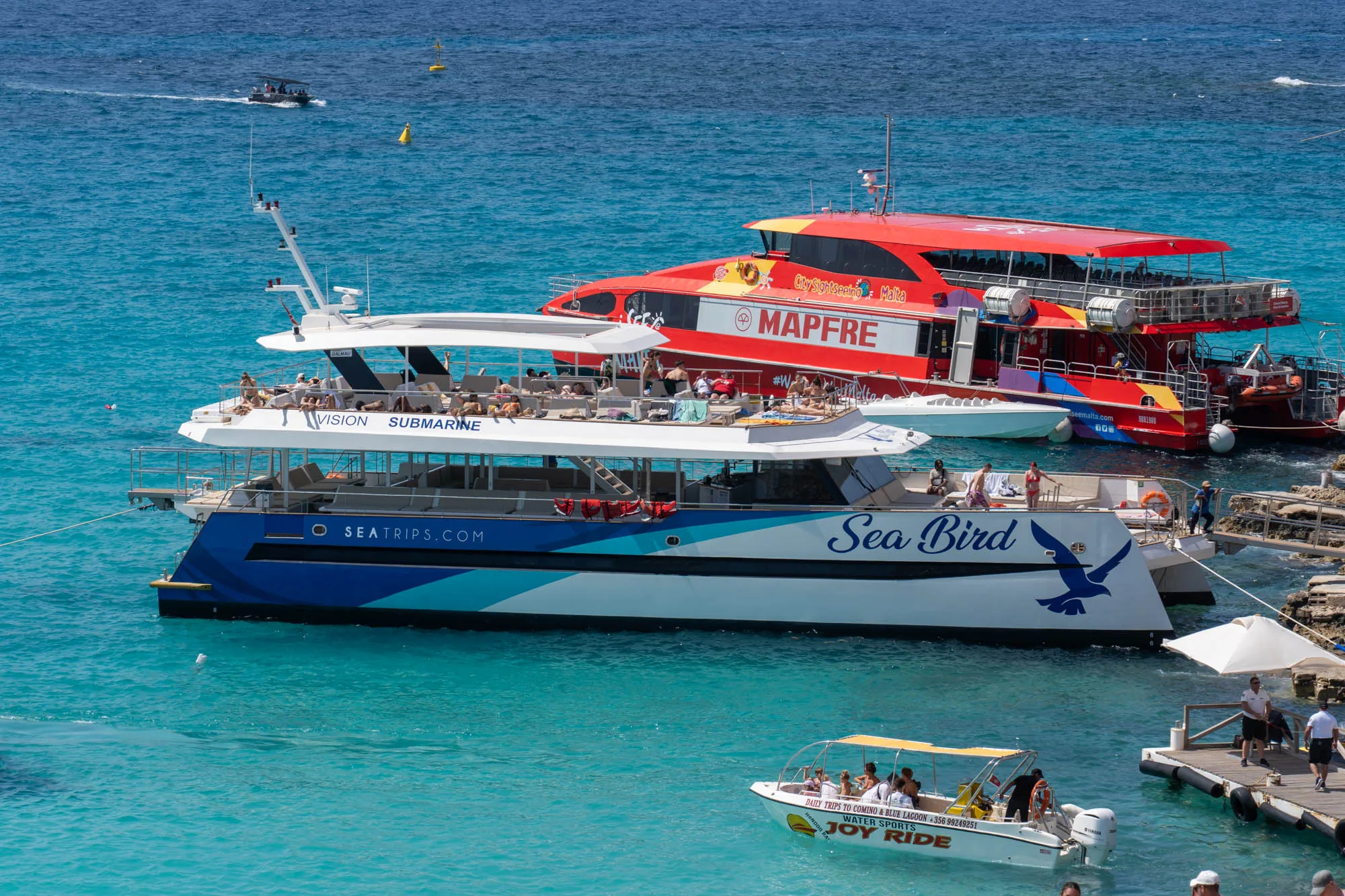 写真はマルタの港で撮影されたもので、複数の観光船が停泊しています。手前には青と白の「Sea Bird」という名前の船が係留されており、船体には大きな青い鳥のイラストが描かれています。その上には「VISION SUBMARINE」という名前の白い船があり、デッキには日光浴をしている人々が多数見られます。奥には鮮やかな赤色の「City Sightseeing Malta」の観光船が停泊しており、MAPFREのロゴが大きく描かれています。さらに遠くには、小さなボートが海を航行している様子が見えます。手前には「JOY RIDE」と書かれた小型ボートも確認できます。水の色は非常に透明度が高く、ターコイズブルーに輝いています。桟橋には人々が歩いているのが見え、全体的に夏の休暇の雰囲気を感じさせる写真です。