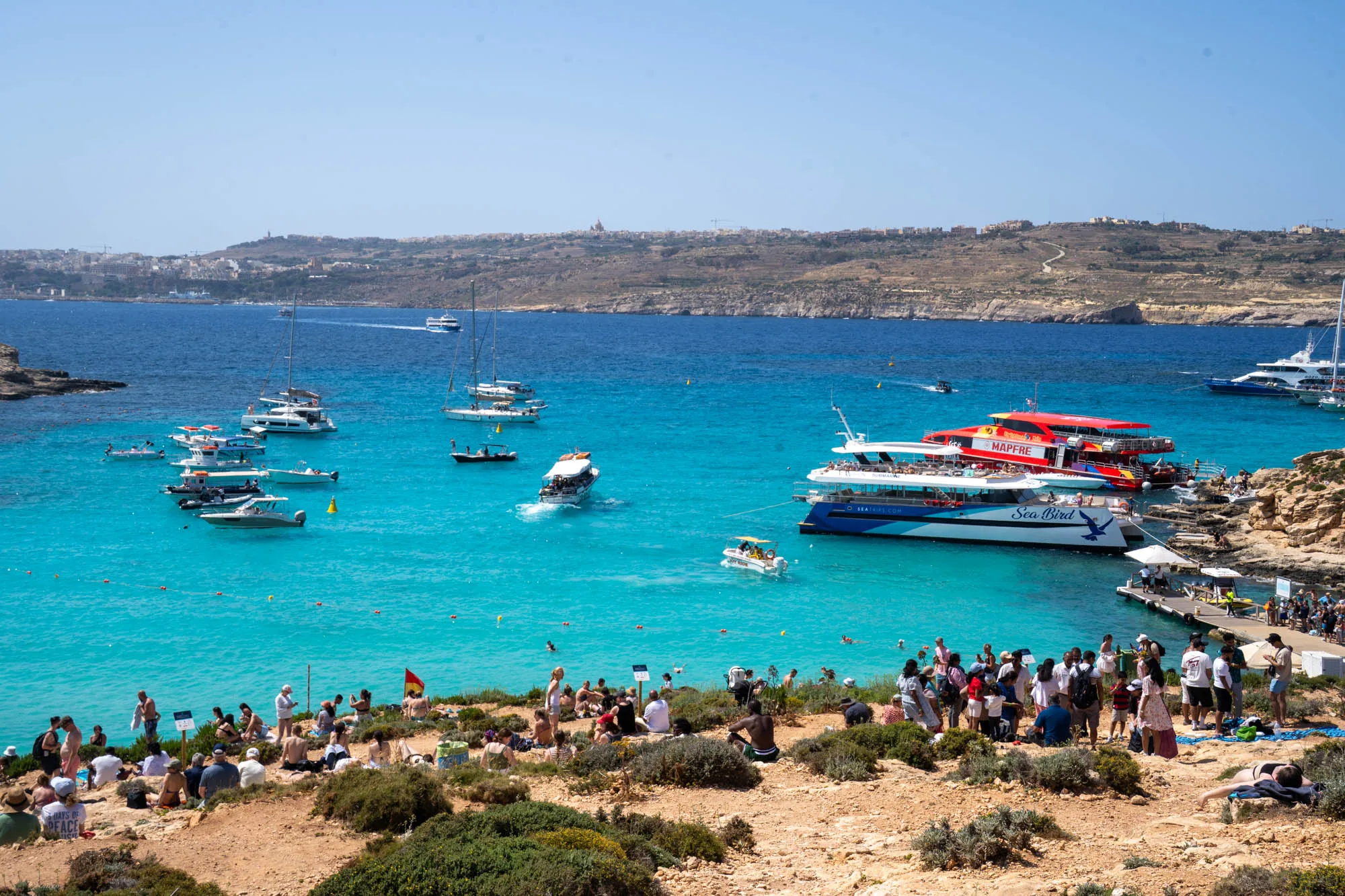 写真は、マルタの海岸線にある美しいビーチの風景を捉えています。ターコイズブルーの透き通った海が広がり、多くのボートやヨットが浮かんでいます。海岸線には、海水浴を楽しむ人々や日光浴をしている人々が多数見られます。岸壁には、人々が乗り降りするための桟橋があり、多くの観光客で賑わっています。遠景には、丘陵地帯の風景が広がり、建物が点在しています。全体的に、明るく活気のある夏の日の様子が伝わってくる写真です。