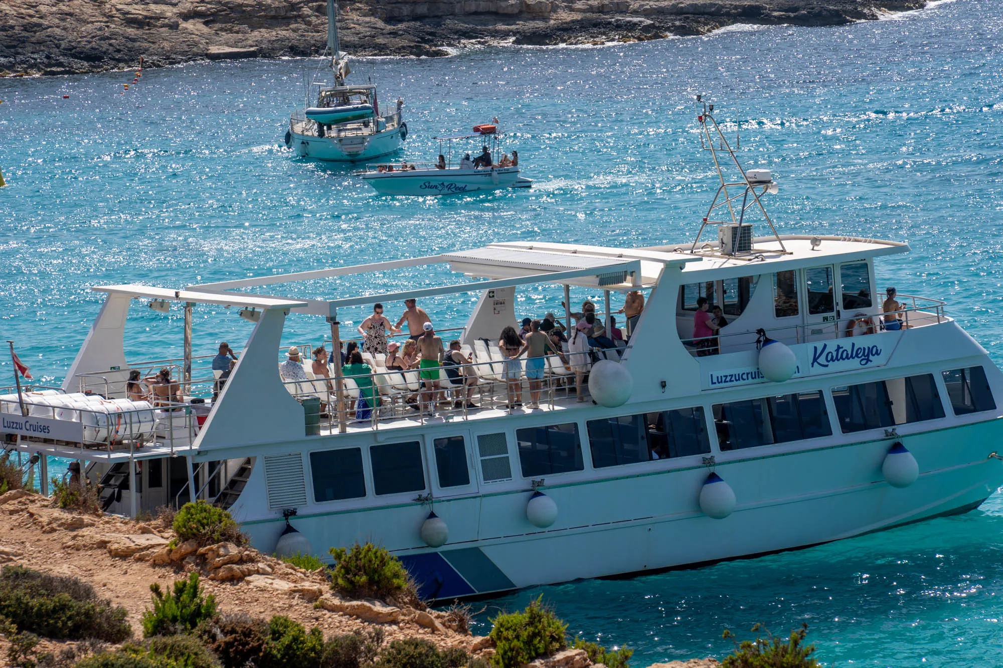 写真には、マルタの美しい海を航行する遊覧船が写っています。船は「Kataleya」と名付けられ、「Luzzu Cruises」という会社が運営しているようです。船上には多くの観光客が乗っており、景色を楽しんだり、日光浴をしたり、リラックスした時間を過ごしている様子が伺えます。背景には、青く輝く海と、遠くに浮かぶ別の船や、岩場が見えます。水の色はターコイズブルーで、太陽の光が水面に反射してきらきらと輝いています。写真全体から、マルタの美しい自然を満喫する人々の楽しい雰囲気を感じることができます。