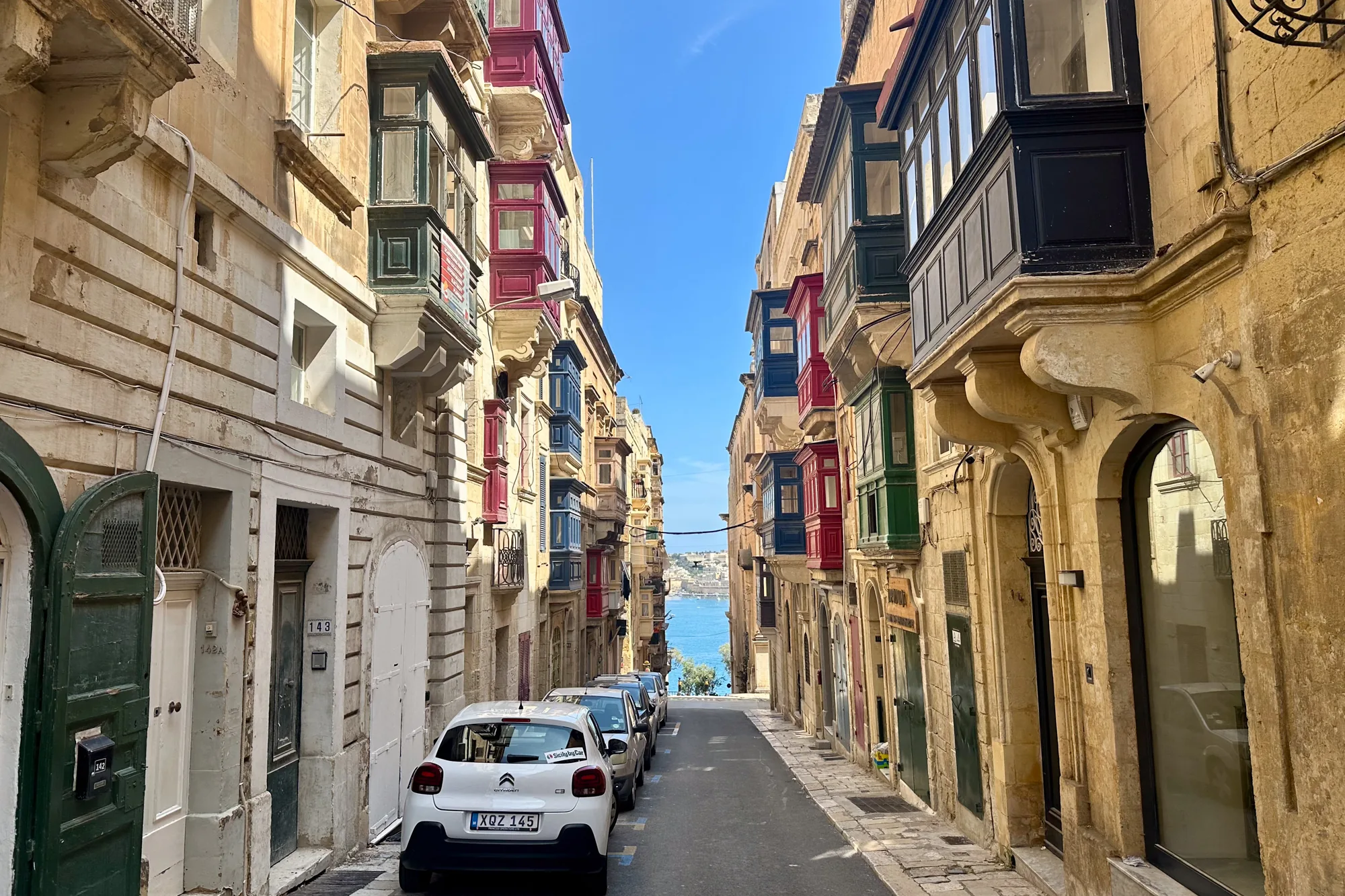写真は、マルタの狭い路地を撮影したものです。路地の両側には、ベージュ色の石造りの建物が立ち並び、赤、青、緑などの鮮やかな色の伝統的な出窓（バルコニー）が特徴的です。路地の奥には、青い海が広がり、遠くの建物がかすかに見えます。路面には車が数台駐車されており、一番手前の白い車が目立ちます。全体的に、マルタの歴史的な街並みの美しさと、地中海の明るい日差しを感じさせる写真です。
