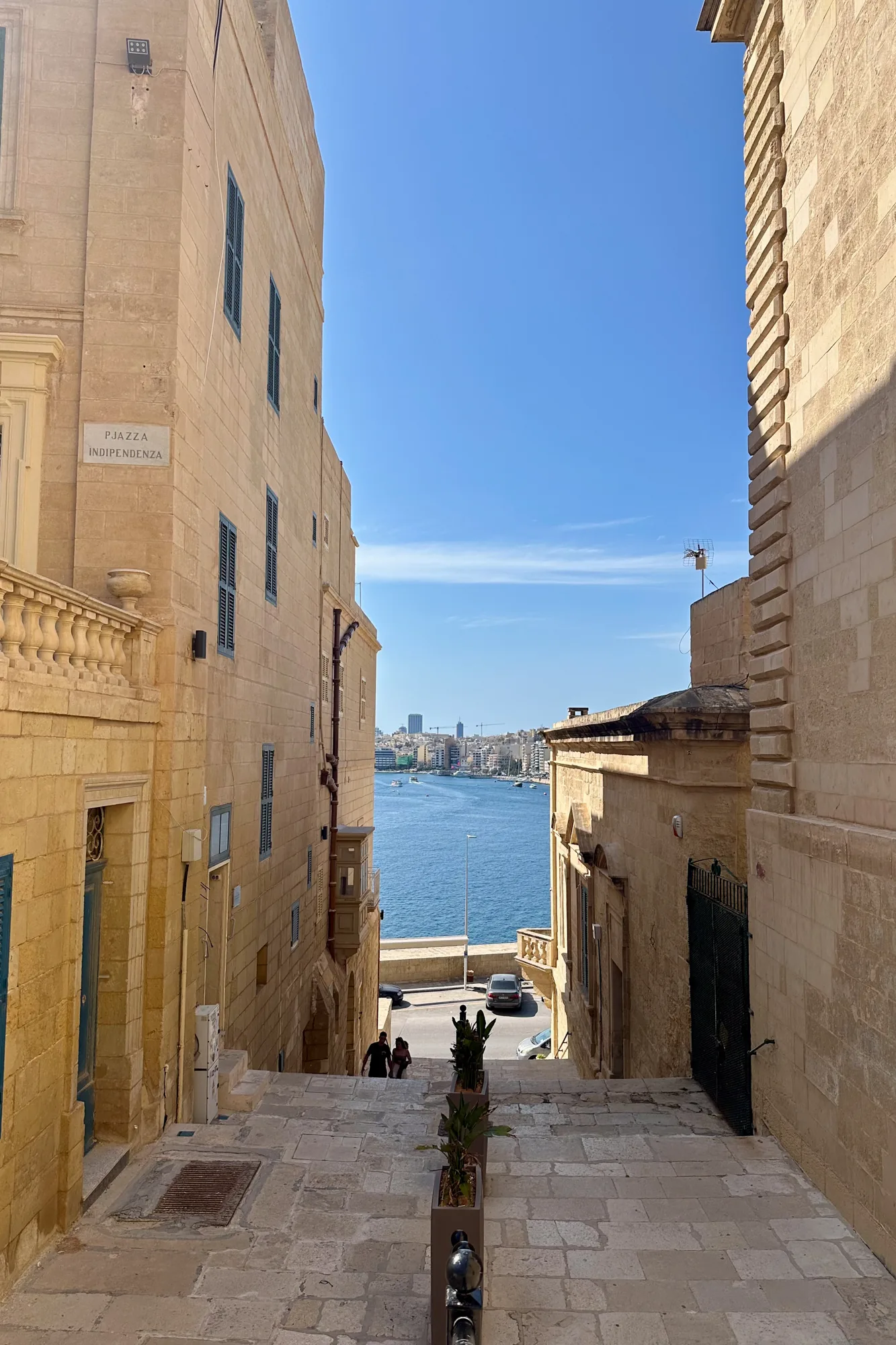 写真はマルタのどこかの場所で撮影されたものです。石畳の階段が画面中央に位置し、その両側を背の高い石造りの建物が挟むように立っています。建物はベージュ色で、古い趣があります。階段を下りた先には青い海が広がり、遠くには都市のスカイラインが見えます。空は晴れており、太陽光が画面全体を照らしています。階段には、人々が歩いている様子が写っています。階段の手すりにはプランターが設置されています。建物の壁には「PJAZZA INDIPENDENZA」と書かれた標識が取り付けられています。写真全体から、マルタの歴史的な街並みと美しい自然が組み合わさった風景を感じられます。