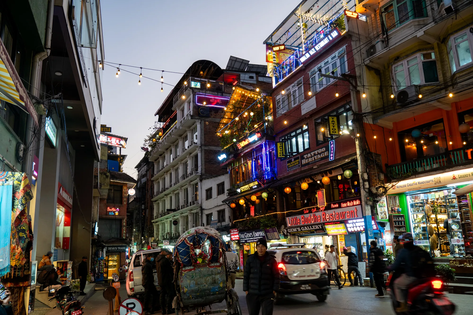 この写真はネパールのカトマンズで撮影された夜の街並みを捉えています。通りには建物が立ち並び、多くはレストランやギャラリーのようです。建物は様々なスタイルで、ネオンサインや装飾的な照明で飾られています。特に「Little Buddha Restaurant」や「TATTOO HERITAGE」といった看板が目を引きます。通りの照明は暖色系の光を放ち、活気ある雰囲気を醸し出しています。人々が行き交い、バイクや車が走っています。手前には人力車があり、観光客の姿も見えます。全体的に、カトマンズの賑やかでエキゾチックな雰囲気が伝わってくる写真です。