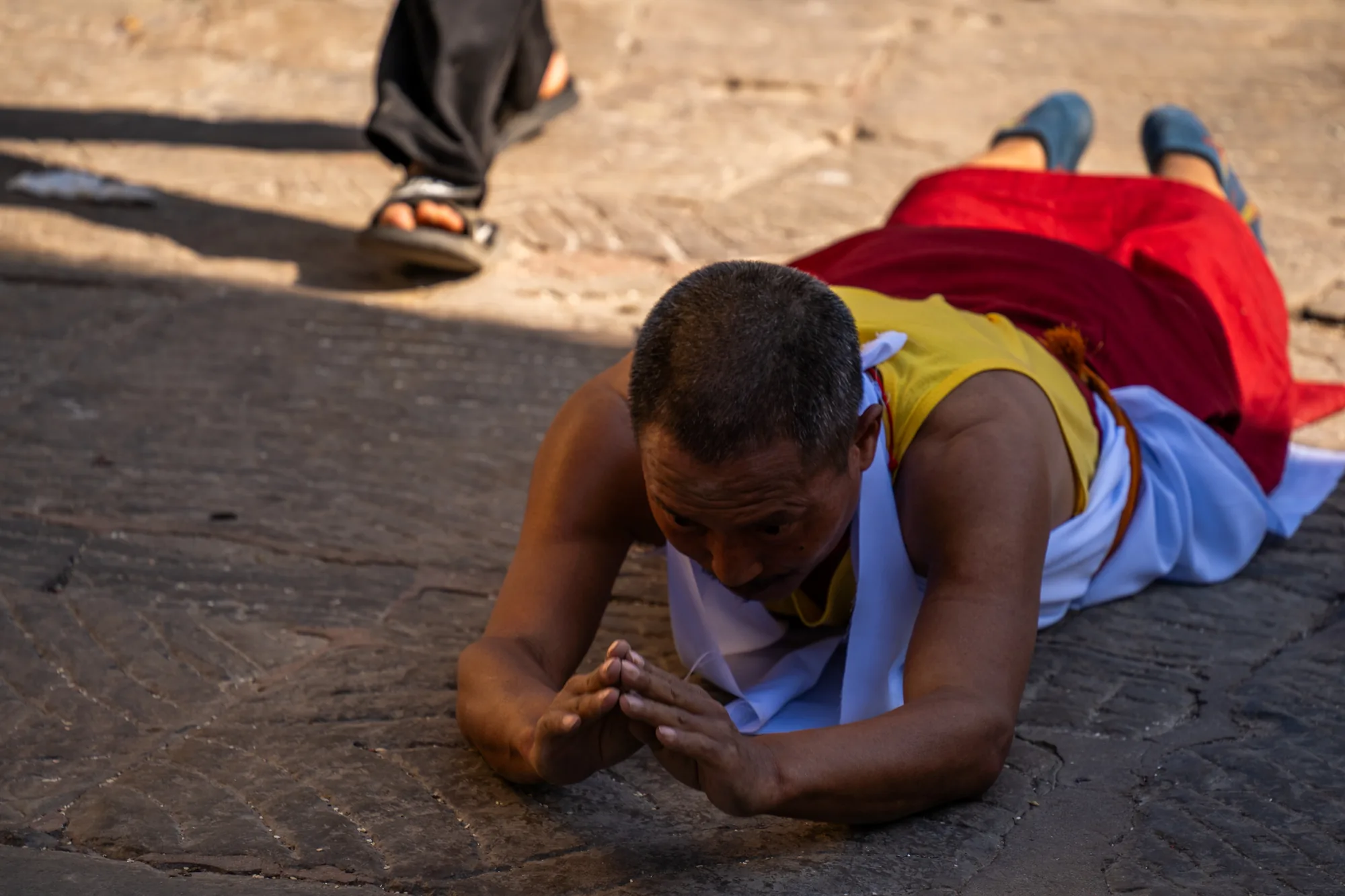 ネパールのカトマンズで撮影された写真には、地面に五体投地をする男性が写っています。男性は黄色と赤色の僧衣を身につけており、首には白い布がかけられています。地面は石畳で、ひび割れなどが見られます。男性は両手を合わせ、祈りを捧げている様子です。光が差し込み、石畳に影を落としています。画面奥には、サンダルを履いた人の足の一部が見えます。全体的に敬虔な雰囲気が感じられます。