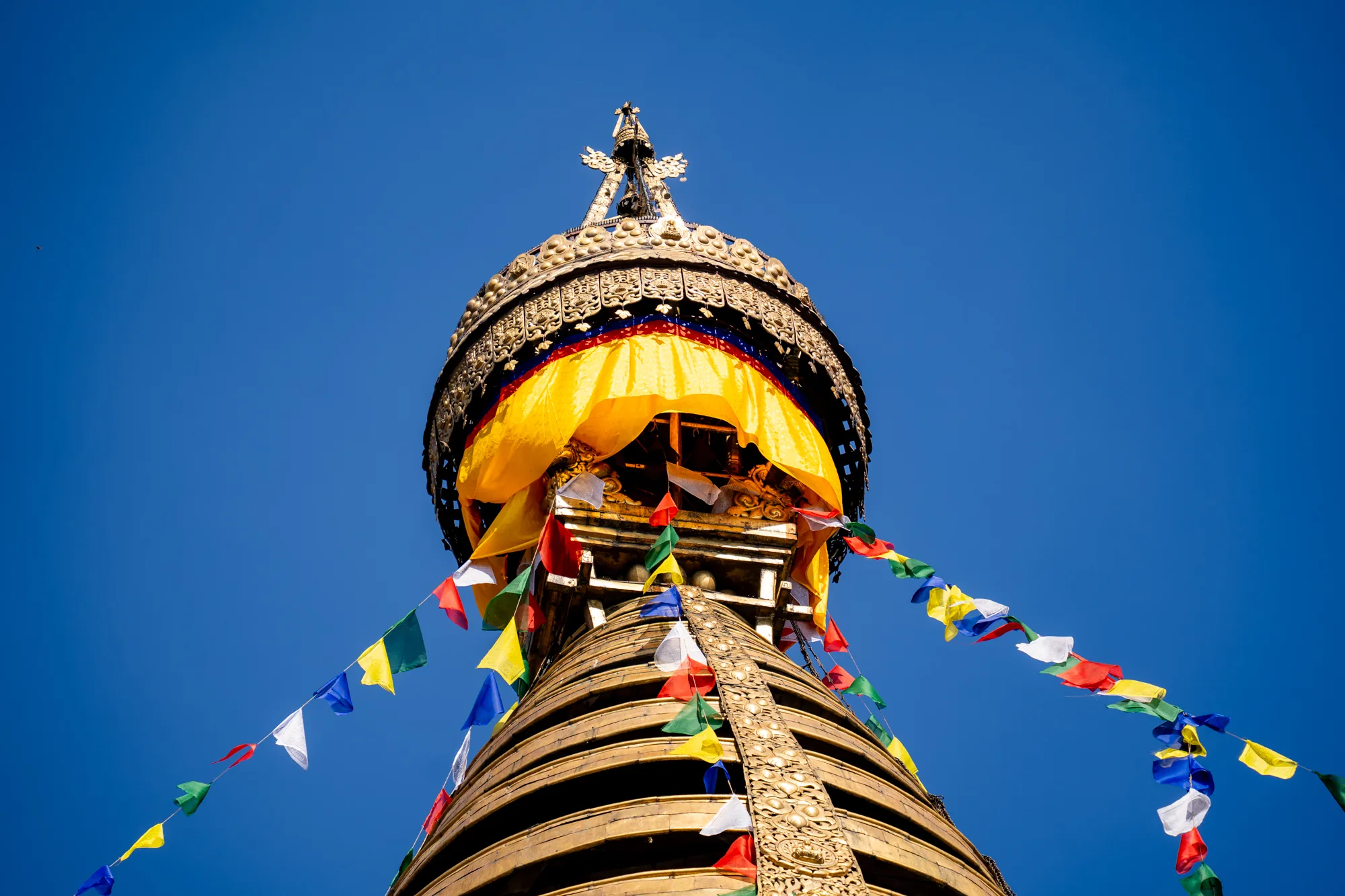 ネパールのカトマンズで撮影された、仏塔の上部をローアングルから見上げた写真です。空は快晴の青色で、仏塔の金色の塔身が鮮やかに映えています。塔身は同心円状に装飾されており、表面には精緻な模様が施されています。塔の上部には、装飾的な傘蓋があり、その下には黄色い布が垂れ下がっています。傘蓋の頂点には、さらに複雑な装飾が施された尖塔がそびえ立っています。仏塔からは、色とりどりのタルチョ（祈祷旗）が風になびいており、全体の雰囲気を華やかにしています。