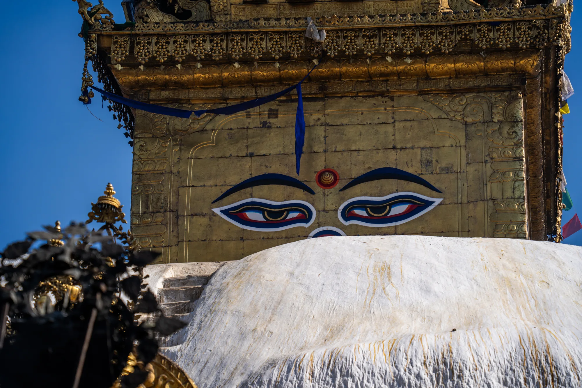 ネパール、カトマンズのスワヤンブナート仏塔の一部を写した写真です。画面中央には、白く塗られた仏塔のドーム状の部分が大きく写っています。ドームの上には、金色の壁がそびえ立ち、その正面には一対の大きな仏陀の目が描かれています。目は、黒、白、青、赤の色で縁取られ、内側の瞳は金色です。眉間には赤い渦巻き模様があり、これは第三の目（智慧の目）を表していると考えられます。金色の壁の上部は、精巧な彫刻で装飾されています。壁の一部には青い布が垂れ下がっています。背景には青空が広がり、仏塔の神聖な雰囲気を際立たせています。写真全体は、鮮やかな色彩と細部へのこだわりが感じられ、ネパールの文化と宗教的な美しさを表現しています。
