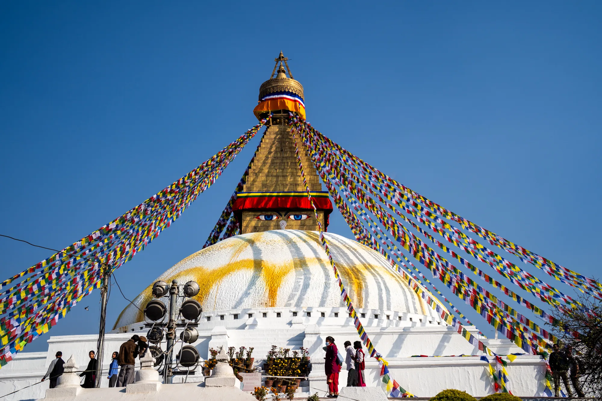 ネパール、カトマンズにあるボダナート（またはボードナート）の壮大な姿を捉えた写真です。鮮やかな青空を背景に、巨大な白いドーム型のストゥーパがそびえ立っています。ストゥーパの頂上には金色の尖塔があり、そこから色とりどりのタルチョ（祈祷旗）が四方へ放射状に伸びています。タルチョは風になびき、活気のある色彩を空に添えています。  ストゥーパの白いドームには、黄色い線状の模様が描かれており、独特の視覚的アクセントになっています。ドームの中央部には、仏陀の知恵の目を象徴する大きな目が描かれており、神秘的な雰囲気を醸し出しています。  ストゥーパの基壇部分には、何人かの人々が写っています。彼らはストゥーパを見上げたり、互いに話をしたりしており、日常の風景の中に溶け込んでいます。 ストゥーパの周囲には緑豊かな植え込みも見られ、自然との調和を感じさせます。全体的に、この写真はボダナートの神聖さと美しさを捉え、ネパールの文化と信仰を伝えています。