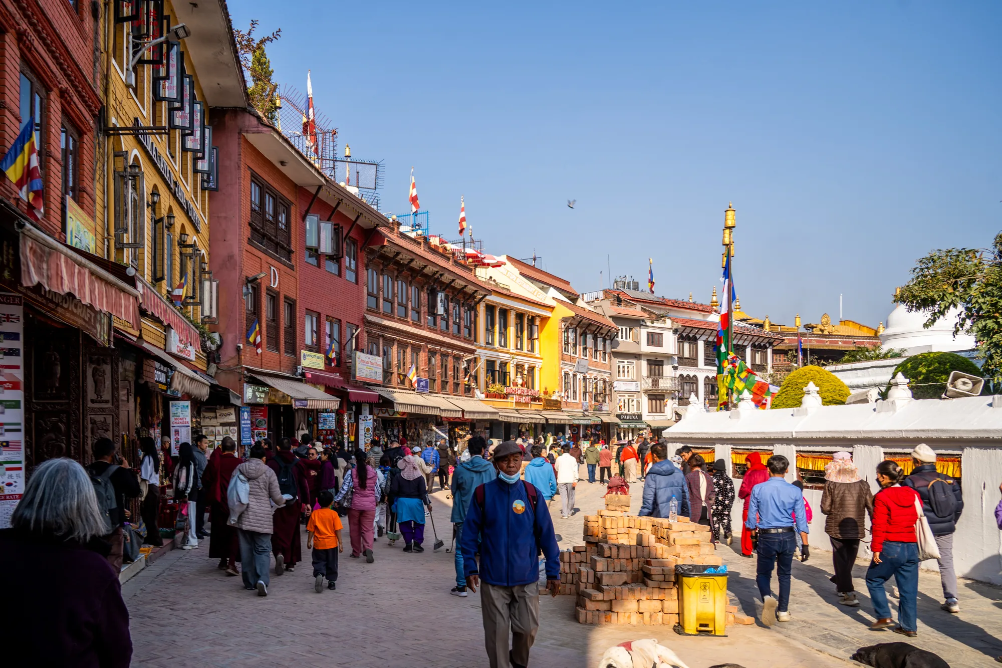 この写真はネパールのカトマンズで撮影された、賑やかな通りの様子を捉えています。道の両側には、色鮮やかな建物が立ち並び、それぞれのお店には、お土産物や日用品などが並んでいるようです。建物の上には、チベット仏教のタルチョのような旗がはためいています。  通りには、多くの人々が行き交っており、観光客だけでなく、地元の人々もいるようです。服装も様々で、民族衣装を着た人や、普段着の人などが見られます。写真全体からは、活気あふれるカトマンズの日常が感じられます。青空が広がり、明るい日差しが差し込む中で、人々のエネルギーと街の色彩が調和した、魅力的な一枚です。