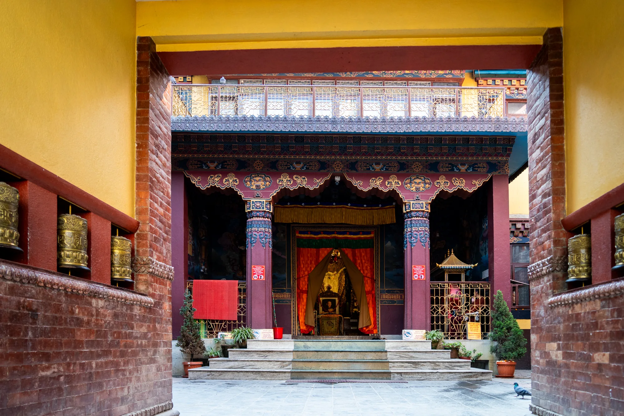 ネパールのカトマンズで撮影された写真です。寺院の境内が写っており、フレームのような構造を通して、奥にある本堂のような建物が見えます。手前は黄色い壁と赤茶色の煉瓦の壁があり、壁にはマニ車が設置されています。奥に見える建物は、赤と青を基調とした装飾が施された柱と屋根を持ち、伝統的な模様が描かれています。階段を上がった先に、金色の仏像のようなものが安置されており、赤いカーテンがかけられています。植物の鉢植えや柵が見え、静寂で神聖な雰囲気が漂っています。手前の床にはハトが１羽写っています。