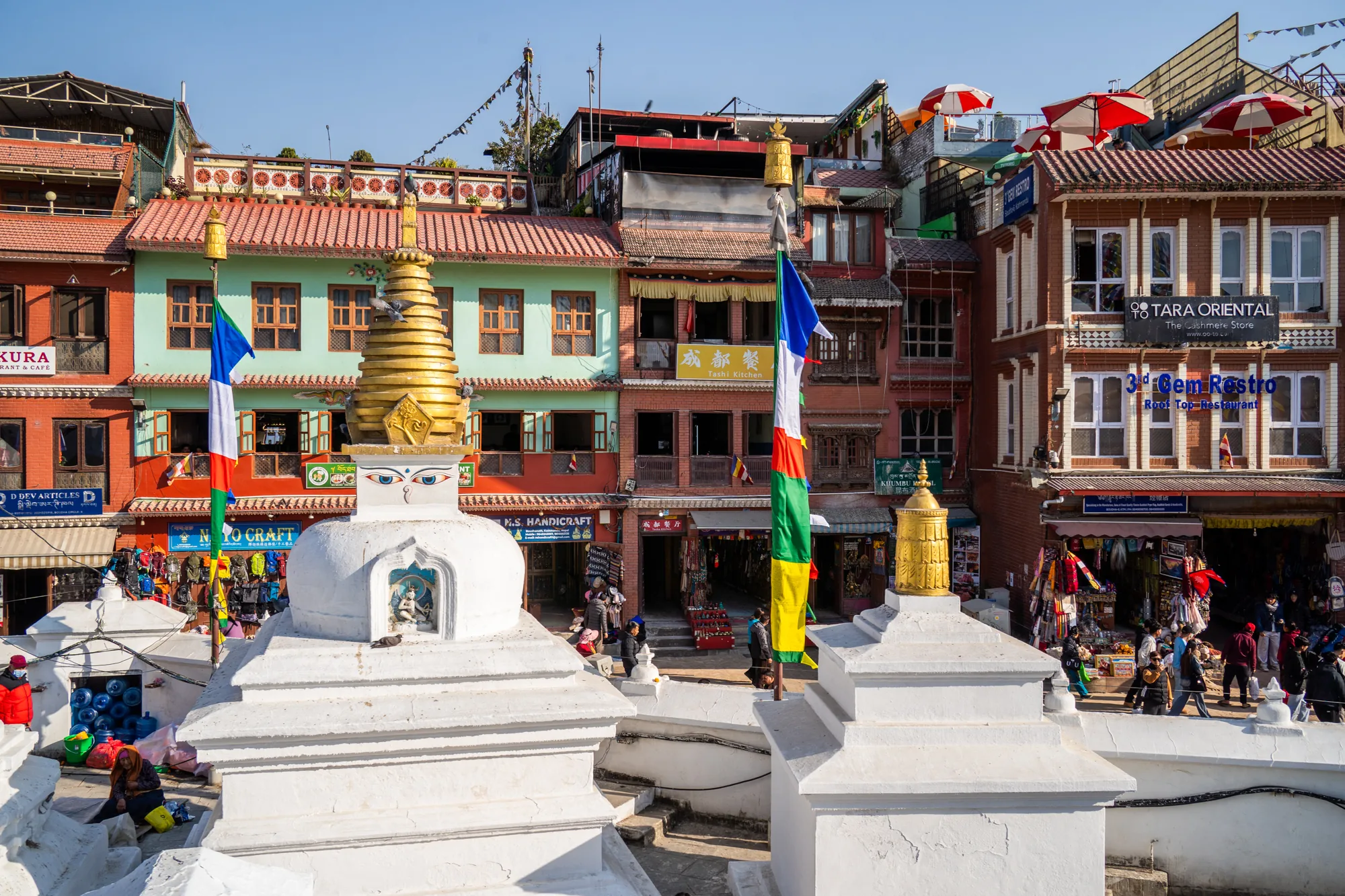ネパールのカトマンズで撮影されたと思われる写真です。手前に白い仏塔（ストゥーパ）が二つ並んでおり、片方の仏塔には金色の装飾が施され、両方の仏塔には仏様の目が描かれています。仏塔の間にはタルチョ（祈祷旗）が立てられています。  背景には、赤、緑、オレンジなどカラフルな建物が密集して建ち並んでおり、ネパールらしい景観が広がっています。建物には、レストランや土産物店など様々な店舗が入っているようです。  通りには多くの人々が行き交い、活気のある様子がうかがえます。「TARA ORIENTAL」や「3d Gem Restro」といった店舗の看板も見えます。屋上にはパラソルが見え、レストランのテラス席のようです。全体的に明るい日差しが差し込み、賑やかでエネルギッシュな雰囲気の写真です。