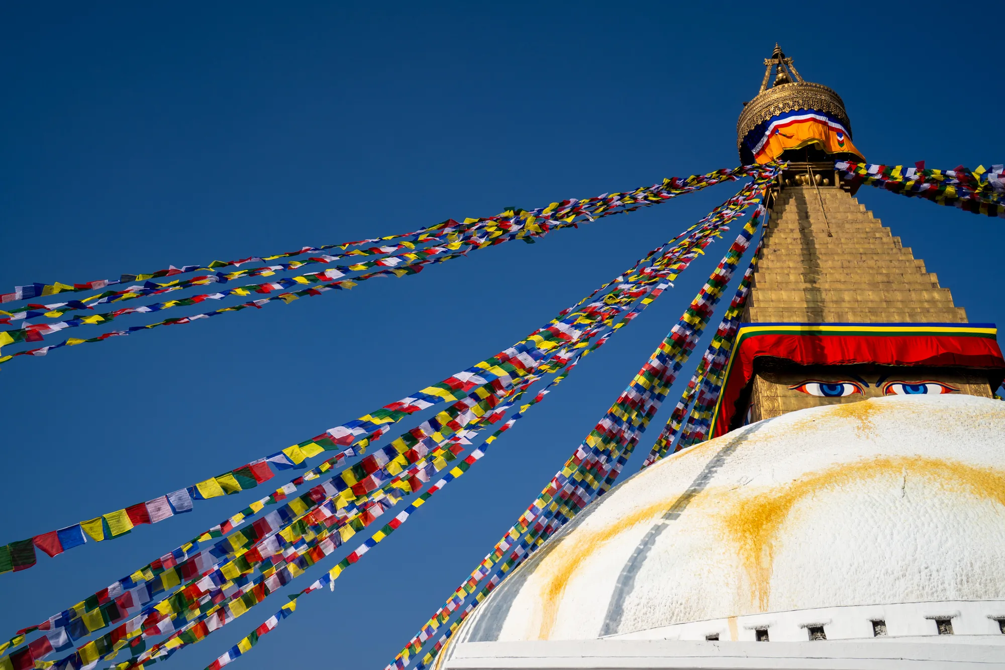 ネパールのカトマンズで撮影されたと思われる、仏塔の写真です。鮮やかな青空を背景に、仏塔の一部が下から見上げるように写っています。仏塔の白いドーム状の部分が大きく写り、その上に、金色の層状の構造物、そして頂上部が確認できます。ドームの上部には、大きな青い目が描かれています。仏塔からは、色とりどりの祈祷旗が四方八方に伸びており、風になびいている様子がうかがえます。祈祷旗は、赤、青、黄、白、緑などの色鮮やかな旗が連なっており、独特の雰囲気を醸し出しています。全体的に、明るく神聖な雰囲気の写真です。