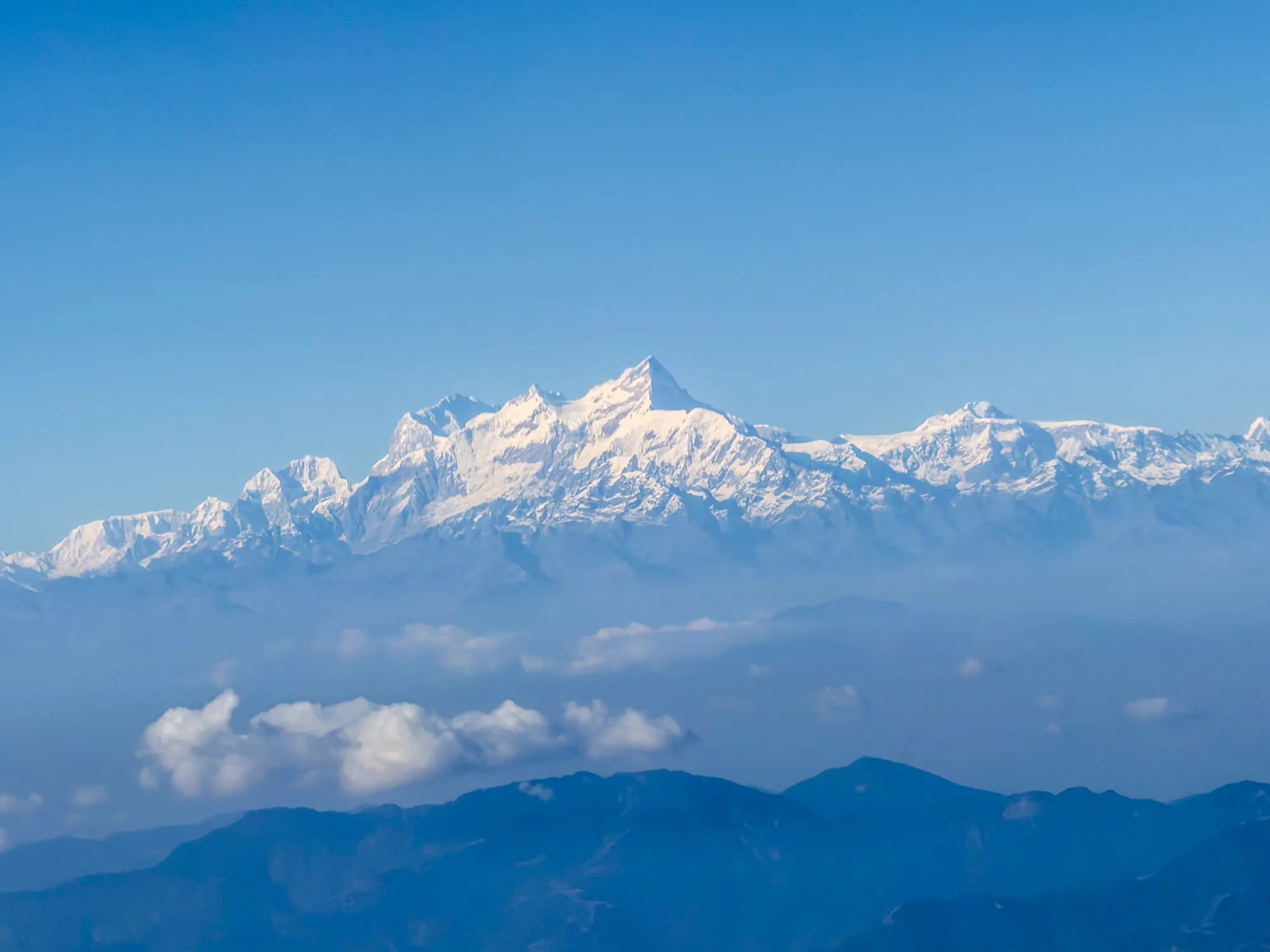 写真はネパールのカトマンズで撮影された、雪を頂く山々の風景です。空は澄み切った青色で、山頂は太陽の光を浴びて白く輝いています。山々の下には雲海が広がり、幻想的な雰囲気を醸し出しています。さらにその下には濃い青色の山並みが連なり、奥行きを感じさせます。全体的に静かで穏やかな、雄大な自然を感じさせる美しい一枚です。