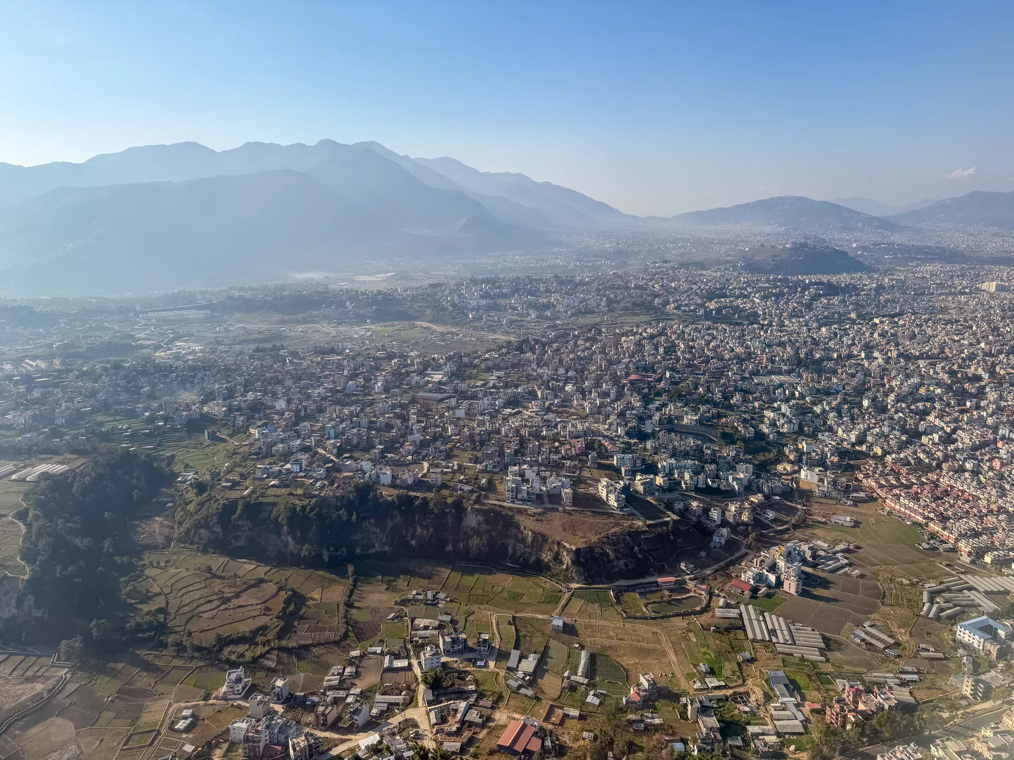 この写真は、ネパールのカトマンズを上空から捉えたものです。広大な市街地が眼下に広がり、家々が密集して建ち並んでいる様子がわかります。その背景には、霞がかった山々が連なり、雄大な自然を感じさせます。市街地の中には、緑豊かな田畑や、段々畑のような地形も見られ、都市と自然が調和している様子が伺えます。空気は少し霞んでいますが、全体的に明るい光に包まれており、穏やかな雰囲気の写真です。