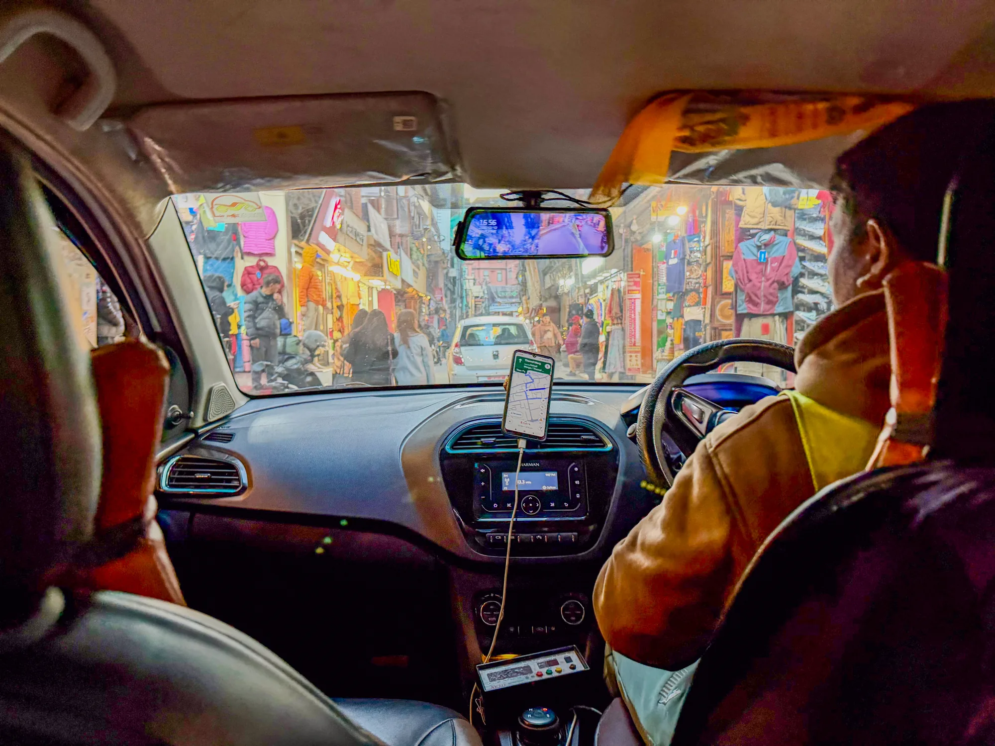 ネパールのカトマンズで撮影された写真です。視点はタクシーの車内からで、運転手の後ろ姿とフロントガラス越しに見える賑やかな街並みが写っています。運転手は黄色のジャケットを着用しており、ハンドルを握っている様子が見えます。タクシーのダッシュボードには、スマートフォンのナビゲーションアプリがセットされ、タクシーメーター、ラジオ、エアコンの操作パネルがあります。フロントガラス越しには、衣料品店や雑貨店が軒を連ねる賑やかな通りが見え、多くの人々が行き交っています。通りの奥には白い車が停車しており、道の狭さが伺えます。写真は、カトマンズの喧騒とした日常風景を切り取ったものです。