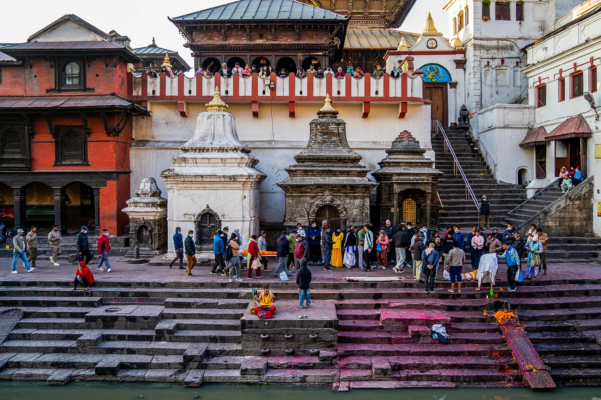 写真はネパールのパシュパティナートで撮影されたもので、聖なるバグマティ川とその周辺の風景を捉えています。川に面した石段には多くの人々がおり、様々な活動をしています。段に座って瞑想していると思われる人物、川のほとりに立って祈りを捧げているような人物、また、集団で歩いている人々など、様々な様子が見られます。石段の一部はピンク色に染まっており、宗教的な儀式が行われた痕跡かもしれません。  背景には、歴史を感じさせる建造物が立ち並んでいます。寺院のような建物、装飾が施された建物、そして、建物のバルコニーから川を見下ろす人々がいます。建造物の様式は独特で、ネパールの伝統的な建築様式を反映しているようです。全体的に、活気があり、信仰心の厚い場所であることが伝わってくる写真です。