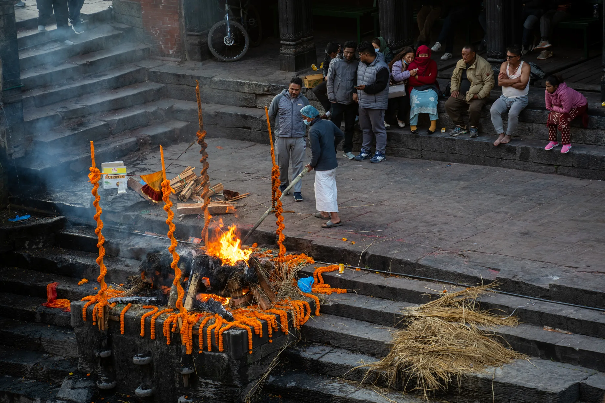 ネパールのパシュパティナートで撮影された写真です。画面中央には、オレンジ色のマリーゴールドの花で飾られた薪の火葬壇があり、炎が勢いよく燃え上がっています。煙が立ち上り、背景の階段の一部を覆っています。火葬壇の前には、白い腰布を巻いた男性が立っており、青い手術帽のようなものを被っています。彼の周りには、数人の男性が立って見守っています。階段には、座って見ている人々が何人か見られます。表情は様々で、悲しみをたたえているように見える人もいます。階段の石の色は灰色で、使い込まれた様子が伺えます。背景には建物の一部や自転車が写っており、場所の雰囲気を伝えています。写真全体から、儀式的で厳粛な雰囲気が感じられます。