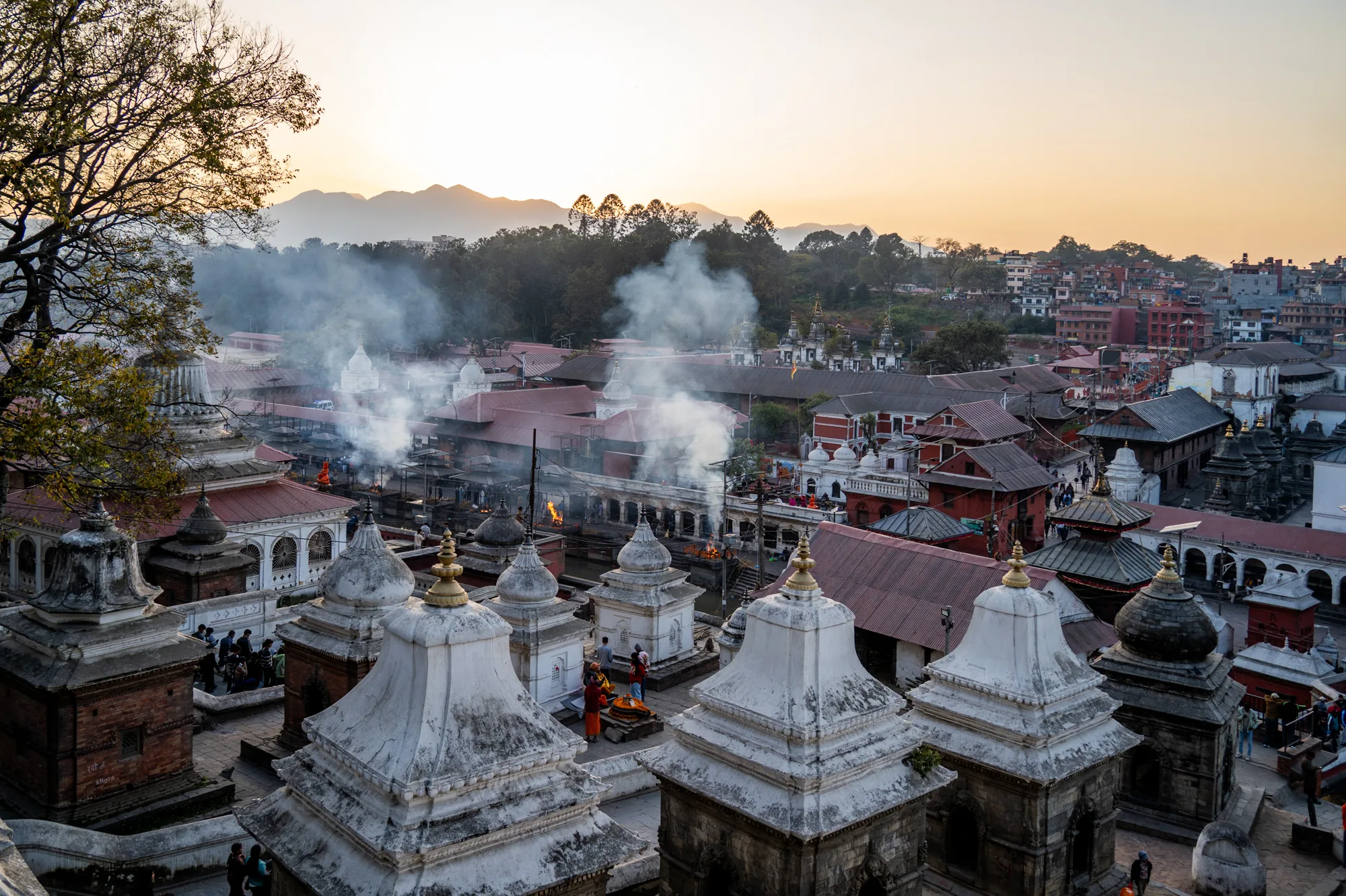 ネパールのパシュパティナートで撮影された写真です。夕焼け時の風景を捉えており、遠景には山々が霞んで見えます。前景には独特の形状をした白い石造りの建造物が複数並んでおり、その間には人々が歩いている様子が伺えます。中央部分には屋根の赤い建物が密集しており、その間から立ち上る煙が印象的です。煙は火葬場から立ち上っているものと思われます。全体的に落ち着いた色調で、神聖で厳かな雰囲気が感じられます。写真全体はやや上からの視点で撮影されており、パシュパティナートの街並みを俯瞰しているようです。文化的な背景と人々の営みが感じられる一枚です。