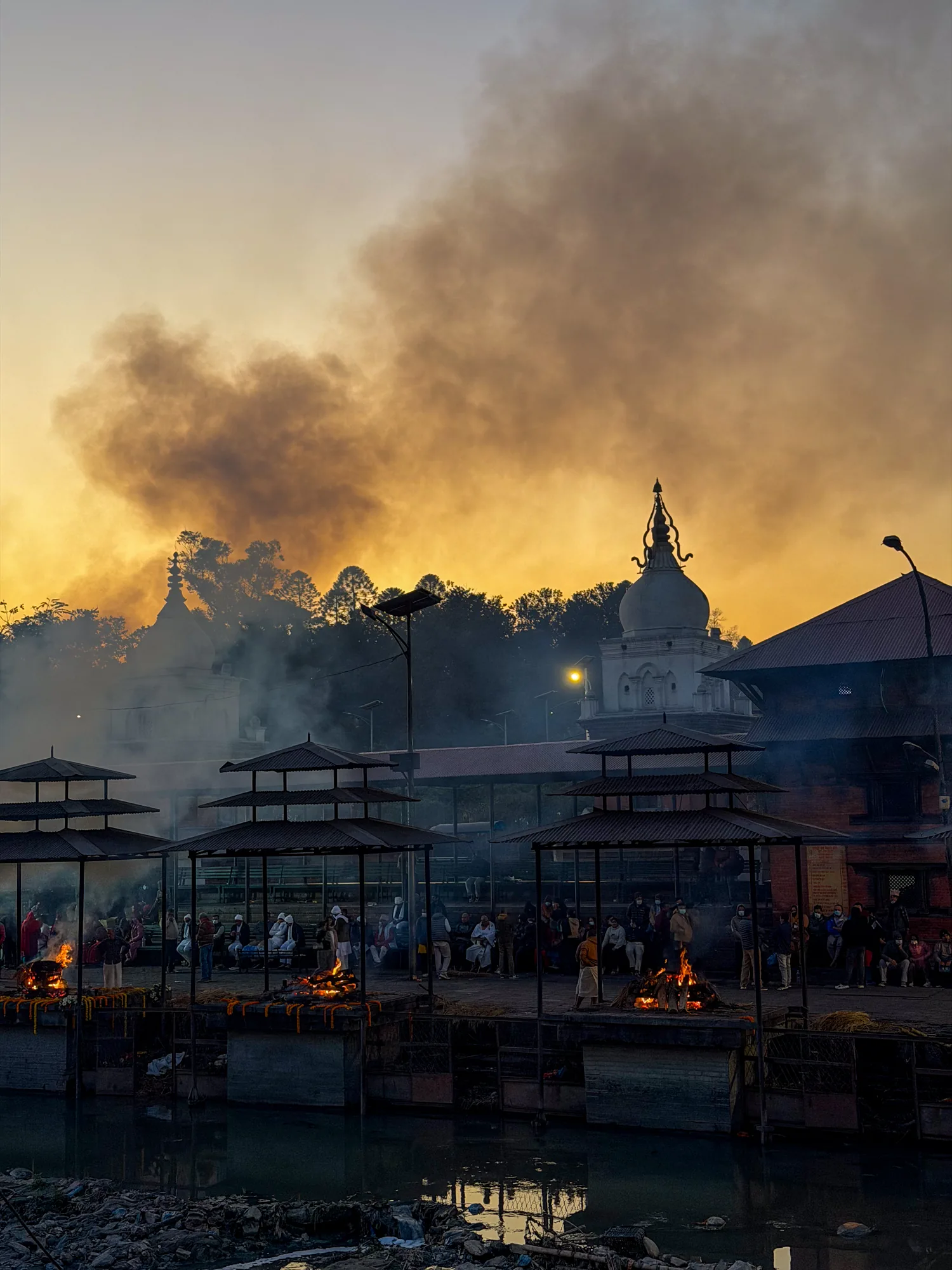 ネパールのパシュパティナートで撮影された写真です。夕暮れ時なのか、空はオレンジ色に染まり、空全体に煙が立ち込めています。手前には、川沿いに設けられた火葬場のような場所が見えます。屋根付きの台がいくつか並び、その上で火が燃え盛っています。人々が周りに集まり、祈りを捧げているようです。川の水面には、燃え上がる炎の光が反射しています。川岸にはゴミのようなものが散乱しており、神聖な場所でありながら、生活感も感じさせる雰囲気です。背景には寺院の建物が見え、シルエットとして浮かび上がっています。全体的に、静かで厳かながらも、生命の終わりを感じさせる、独特な雰囲気の写真です。