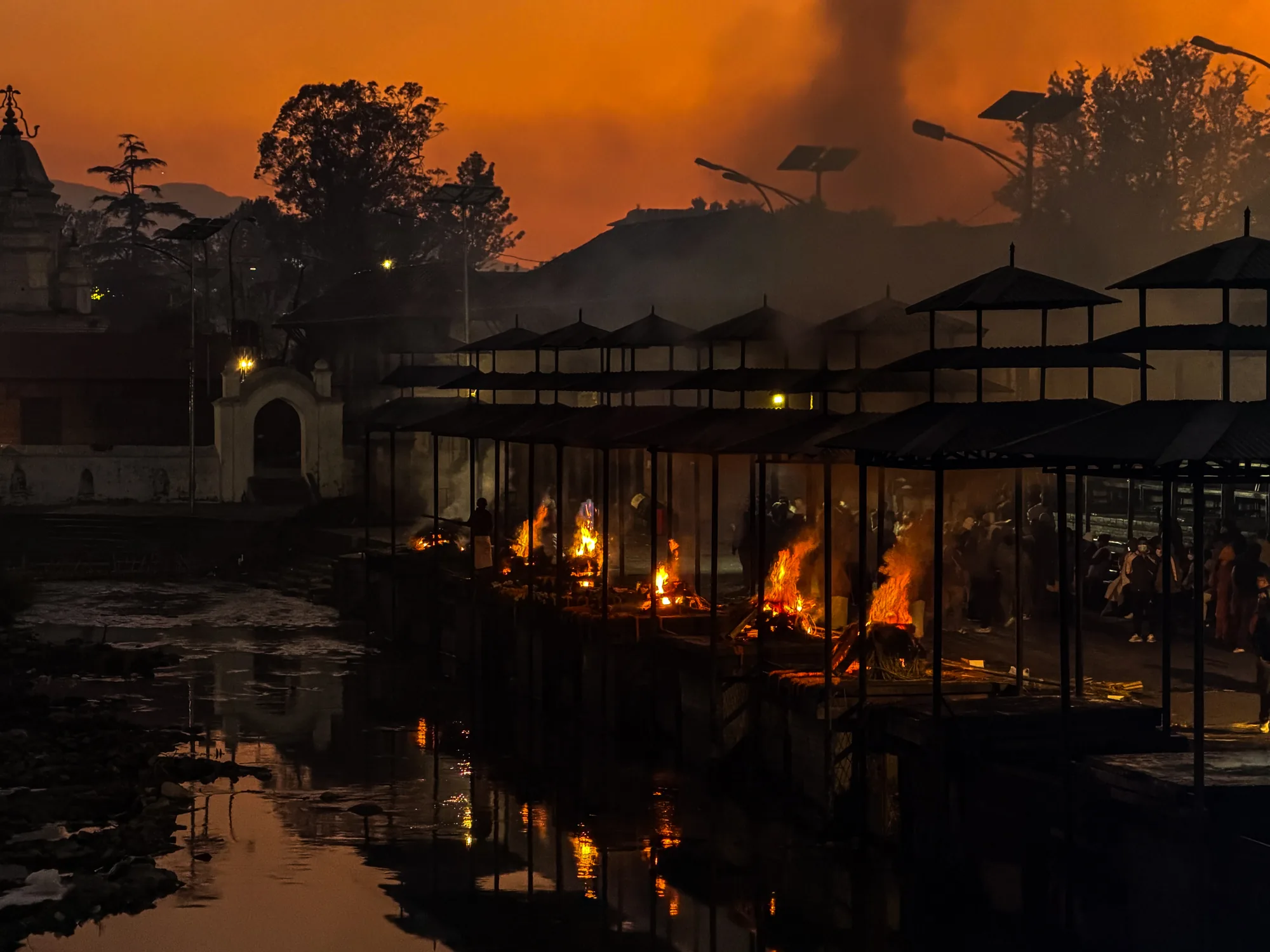 ネパールのパシュパティナートで撮影された写真です。夕暮れ時のような空がオレンジ色に染まり、煙が立ち込めています。画面中央には、川沿いに並んだ火葬場で、いくつもの炎が激しく燃え上がっています。炎は川面に映り込み、揺らめく光を放っています。火葬場の屋根はシルエットとして黒く浮かび上がり、その奥には木々や建物が見えます。橋のような場所には人々が集まり、火葬の様子を見守っているようです。全体的に厳かで、少し神秘的な雰囲気が漂っています。生の終焉と再生の場所であるパシュパティナートの、独特な空気が感じられます。