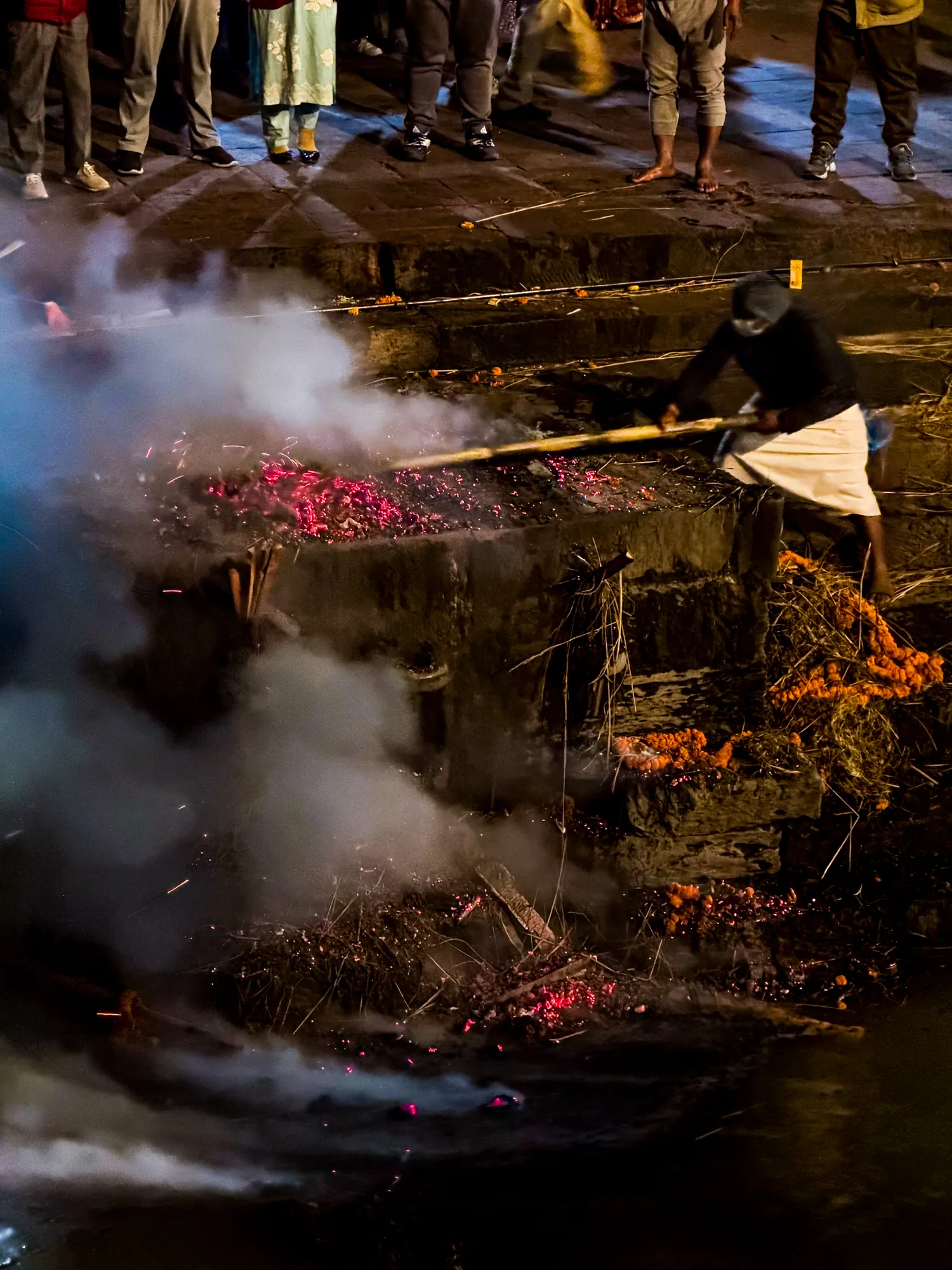 ネパールのパシュパティナートで撮影された写真です。夜の暗闇の中、火葬の様子が写っています。中央には石造りの祭壇のようなものがあり、その上で激しく火が燃え盛っています。燃え盛る炎は赤く、そこから立ち上る煙が周囲を覆っています。祭壇の前には、燃え残った薪のようなものや、燃え尽きた花飾りが散らばっています。祭壇に寄り添うようにして、白い腰布を身に着けた男性が、長い棒を使って火をかき混ぜている姿が見えます。顔は覆われていて判別できません。祭壇の奥には、大勢の人々が立って見守っているようです。足元しか見えませんが、その様子から、静かに故人を偲んでいる様子が伝わってきます。全体的に暗く、煙と炎の赤色が際立っていることから、厳粛で神秘的な雰囲気が漂う一枚です。