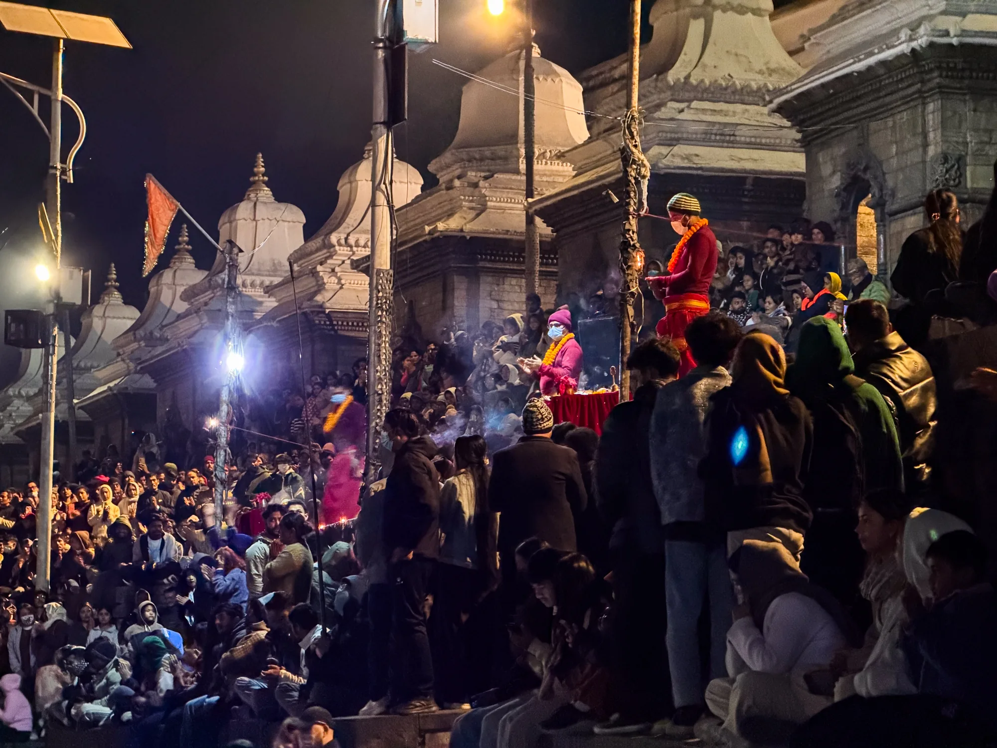 ネパールのパシュパティナートで撮影された夜の写真です。多くの人々が集まっており、お祭りのような雰囲気です。石造りの建物の前には大勢の人が座って何かを見ているようです。建物の上や、手前に赤い衣装を身につけた人物が立っており、儀式のようなことを行っているように見えます。多くの人がマスクを着用しており、コロナ禍であることを示唆しています。夜間のため全体的に暗いですが、建物を照らすライトや、人々が持つ明かりによって、幻想的な雰囲気が醸し出されています。