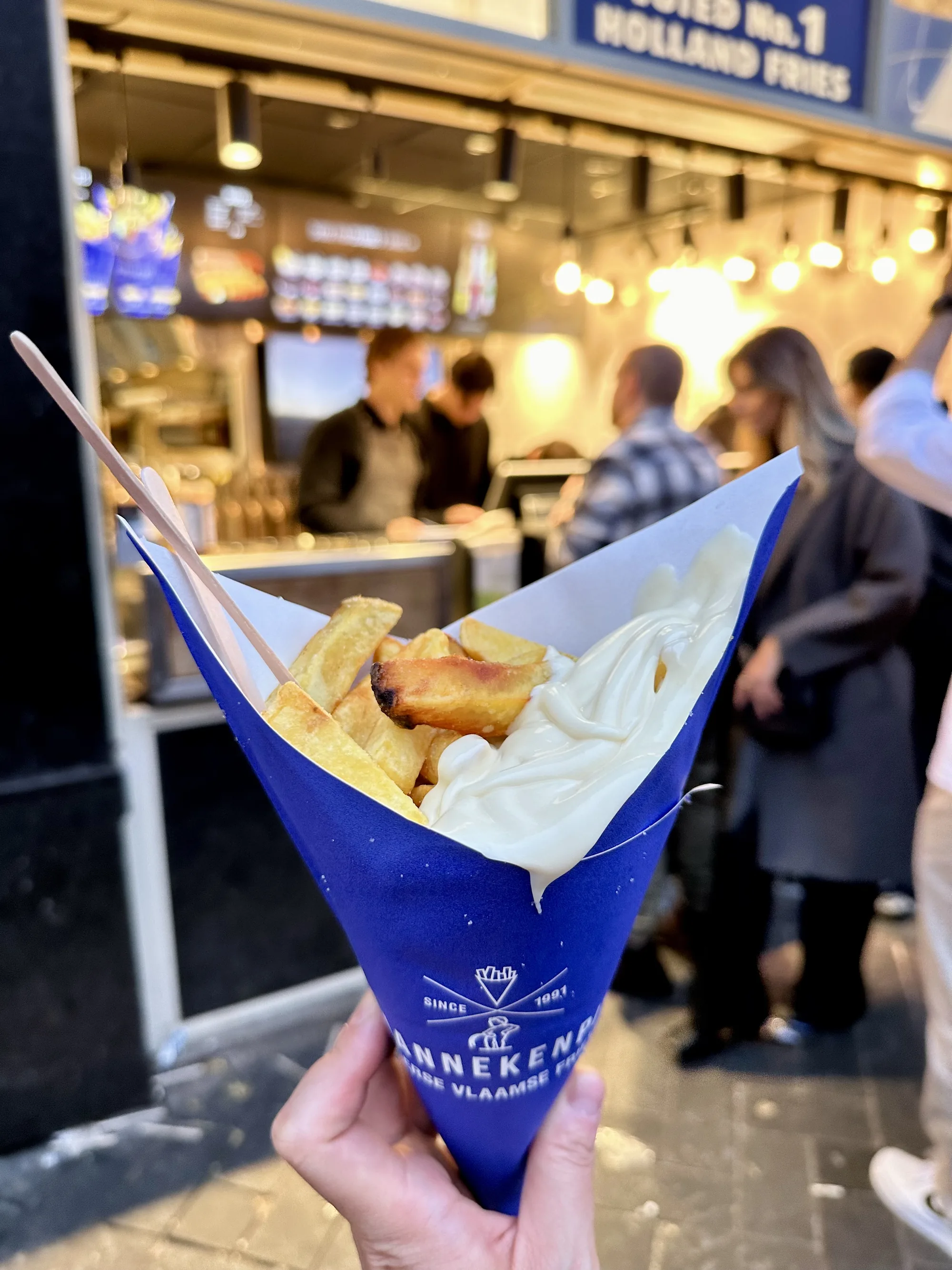オランダ・アムステルダムで撮影された写真です。手には、青色の円錐形の容器に入ったフライドポテトが握られています。ポテトは揚げたてで、表面はカリッとしており、中はホクホクとしていそうです。上にはたっぷりの白いソース、おそらくマヨネーズがかかっています。容器には「Mannekenpis Vlaamse Fries」という文字とロゴがプリントされており、1991年から続くフライドポテト店のものであることがわかります。背景には、お店のカウンターがあり、店員が何人か働いている様子が見えます。また、並んでいる人々の姿も写っており、人気店であることが伺えます。全体的に、アムステルダムの街角でフライドポテトを楽しむ様子を捉えた、食欲をそそる一枚です。