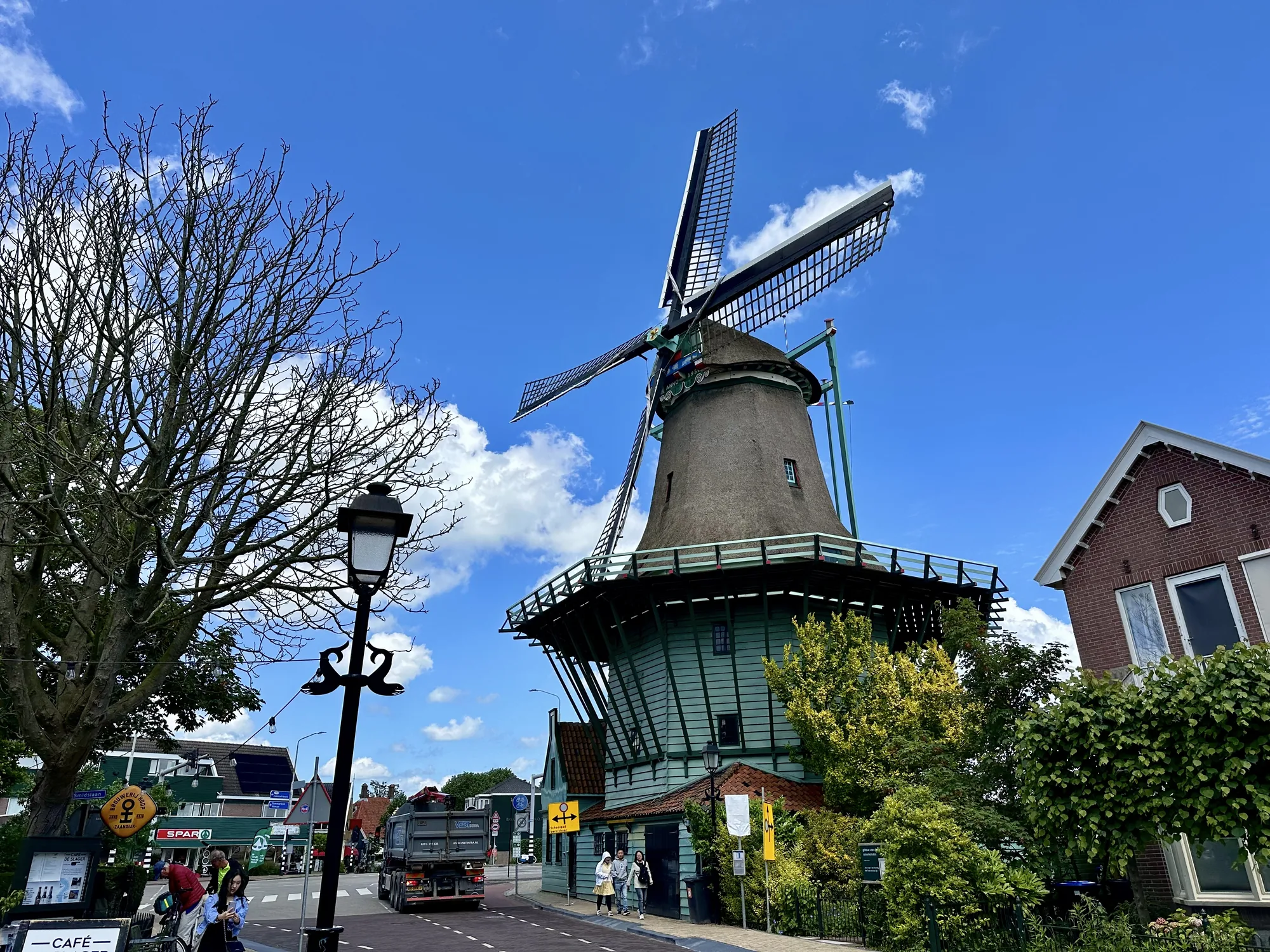 オランダのアムステルダム近郊の風景写真です。青空の下、画面中央に大きく風車が写っています。風車は木造で、下部は緑色、上部は藁葺き屋根のようです。風車の羽は大きく、特徴的な形をしています。風車の周辺は街並みで、左手には葉の落ちた木と、街灯、カフェの看板が見えます。カフェの前には数人の人がおり、右側にはレンガ造りの家が写っています。道路にはトラックが走っており、交通標識なども見えます。空は青く、白い雲が浮かんでおり、全体的に穏やかで美しい風景です。