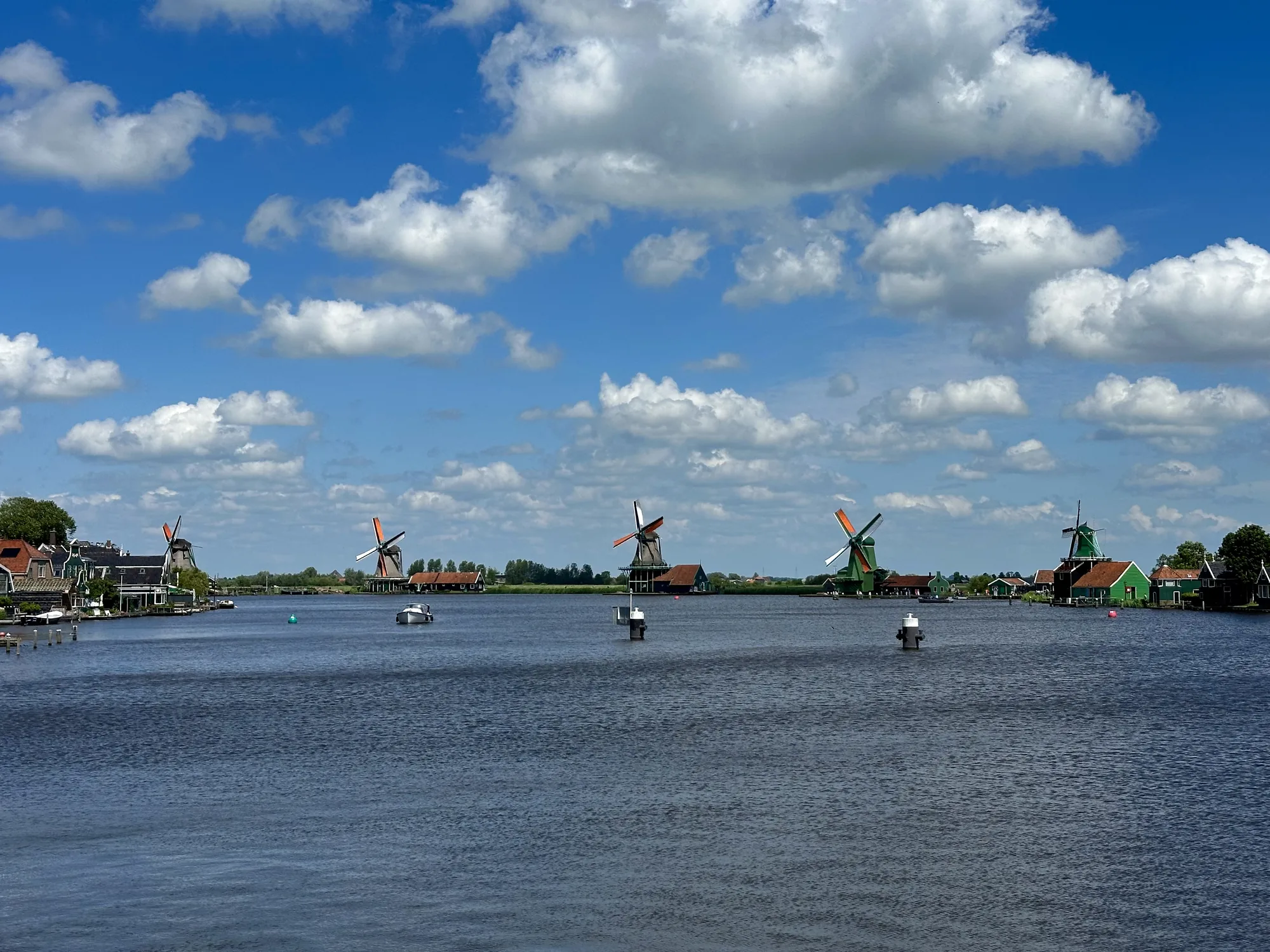 この写真は、オランダのアムステルダム近郊の風景を捉えたものです。前景には穏やかな水面が広がり、水路を示す標識が点在しています。水面には、いくつかのボートが浮かんでいます。水路の向こうには、伝統的なオランダの木造家屋が並び、その背景には特徴的な風車が複数立っています。風車はそれぞれ異なる色合いで、風景に彩りを添えています。空には、白い雲が浮かび、青い空とのコントラストが美しいです。全体的に、のどかで平和な雰囲気が漂っており、オランダの典型的な風景を象徴するような写真です。