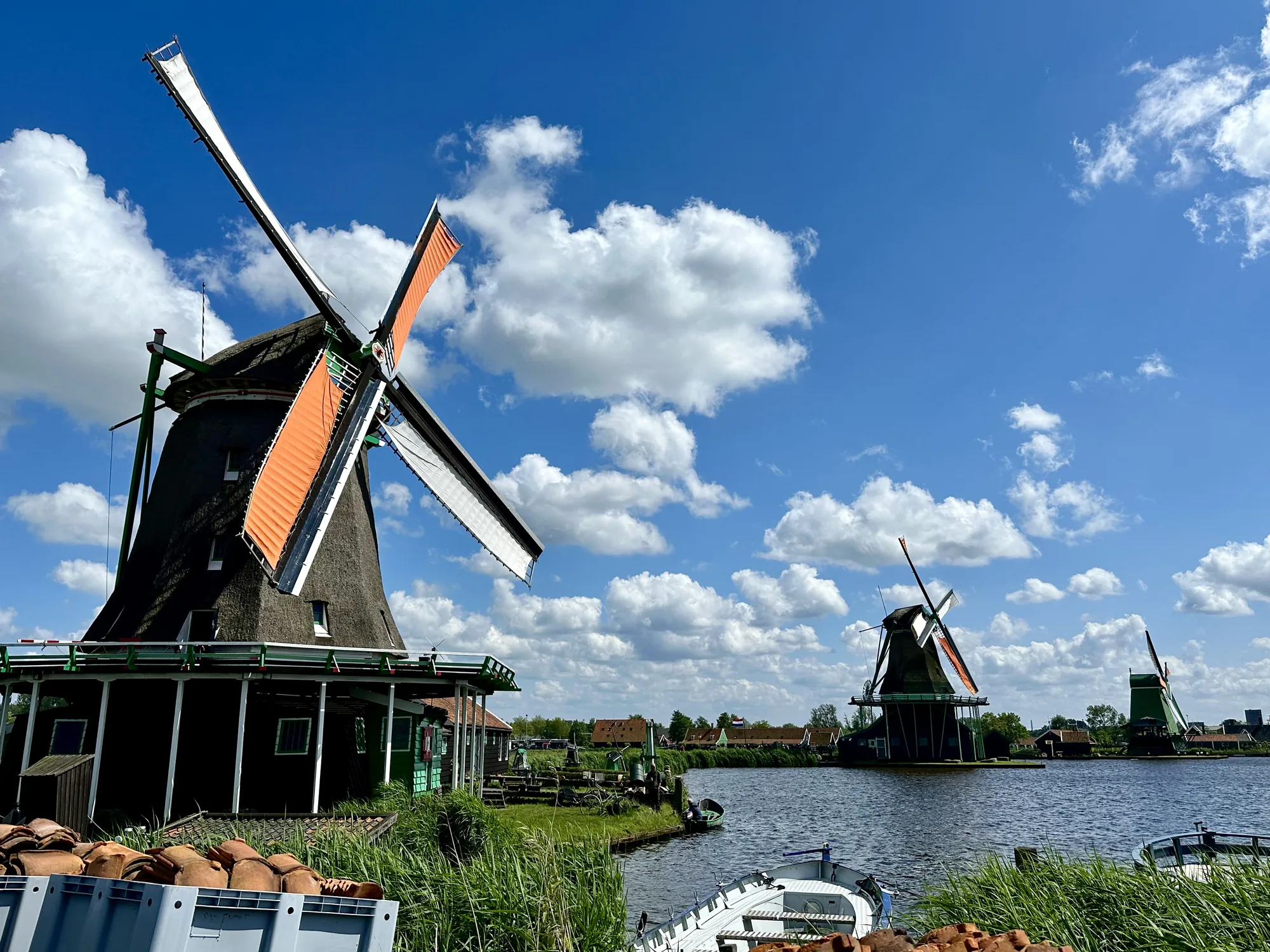 写真は、オランダの典型的な風景である風車が複数写っています。手前には黒っぽく見える風車が立っており、オレンジと白の羽根が印象的です。空は青く、白い雲が点在しており、穏やかな天候であることがわかります。風車の周囲は水辺になっており、緑の草が生い茂っています。奥にも複数の風車が見え、伝統的なオランダの風景が広がっています。手前にはボートや屋根瓦のようなものも写っており、生活感も感じられます。全体的に穏やかで美しい風景写真です。アムステルダム近郊のZaanse Schansのような場所であると思われます。