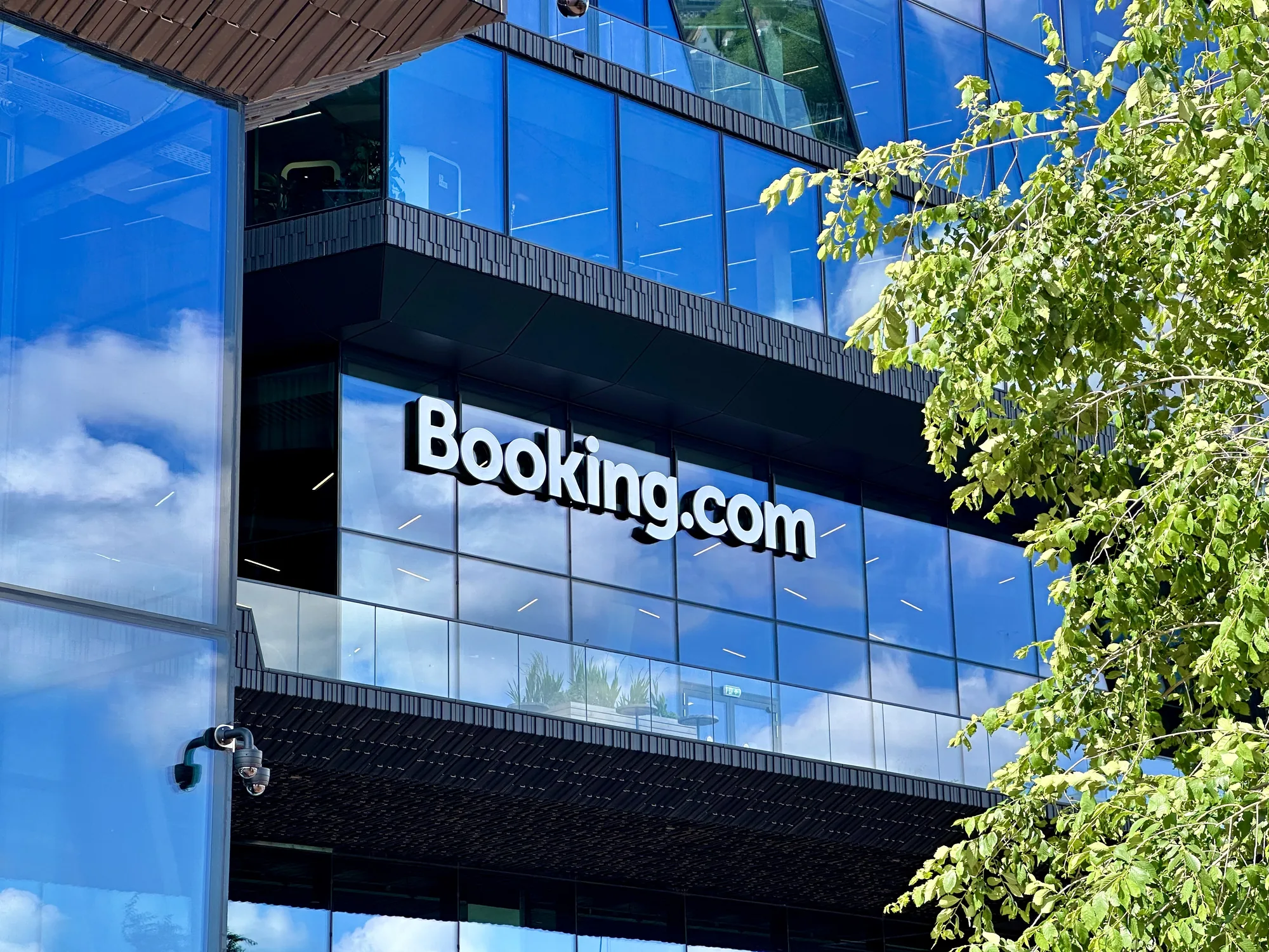 これは、オランダのアムステルダムで撮影されたBooking.comの本社の外観の写真です。モダンな建築様式で、ガラス張りの壁面が特徴的です。壁面にはっきりと「Booking.com」のロゴが表示されています。ガラスには周囲の空の雲が反射しており、建物の存在感を際立たせています。写真の右側には緑豊かな木の枝が写り込んでおり、都市の風景の中に自然の要素が取り入れられています。また、建物の角にはセキュリティカメラが設置されているのが確認できます。全体的に、企業の本社としての洗練された印象と、アムステルダムの晴れた日の雰囲気が伝わってくる写真です。