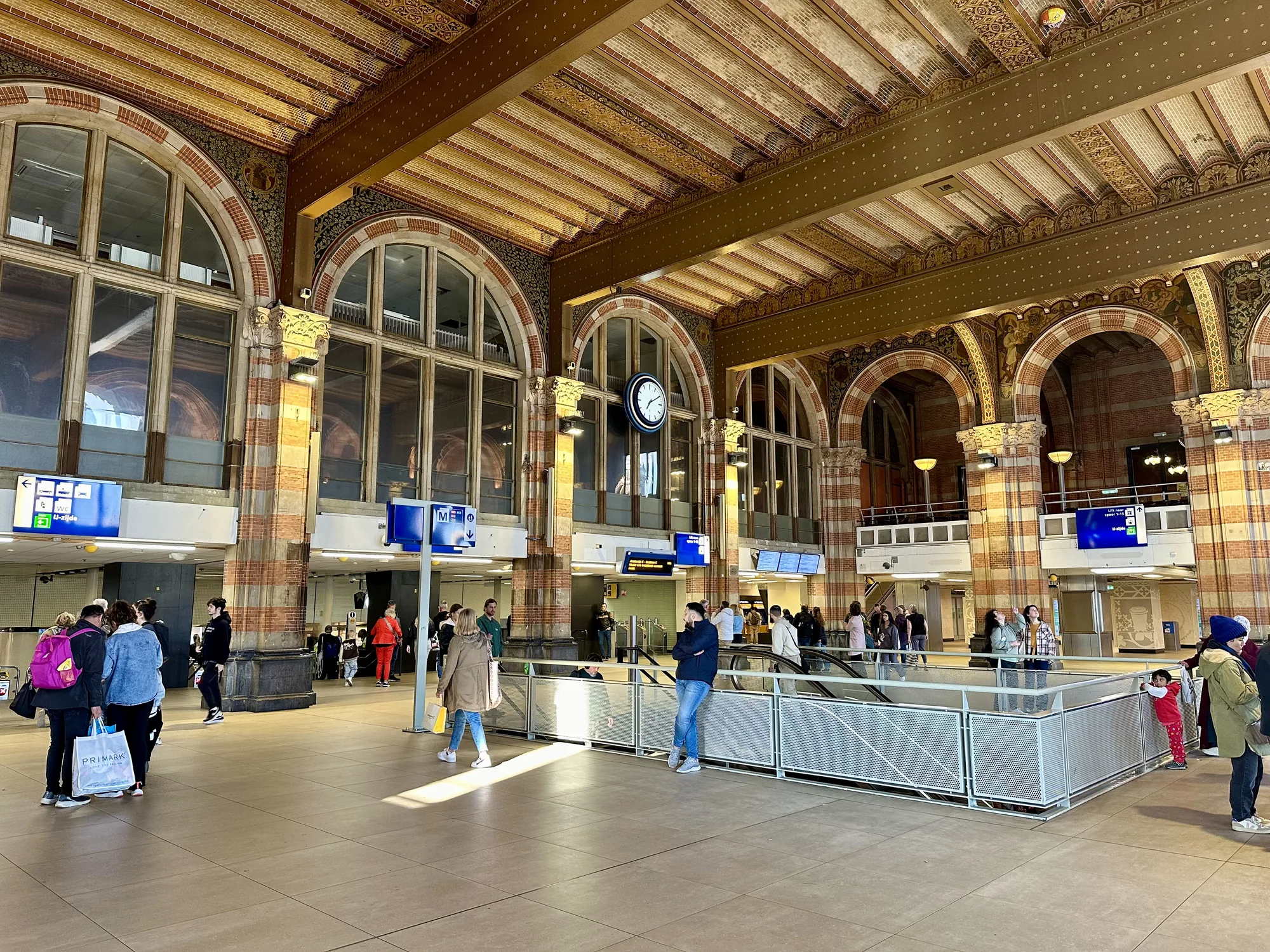 この写真はオランダのアムステルダムにある中央駅の内部を写したものです。天井はレンガと装飾が施され、歴史を感じさせる美しい内装です。大きなアーチ型の窓が並び、自然光が差し込んでいます。構内には人々が行き交い、エスカレーターが見えます。駅の案内表示や時計などが設置されており、多くの人が利用する活気のある場所であることが伺えます。レンガ造りの柱や壁の装飾は非常に細かく、歴史的な建造物としての風格を感じさせます。全体的に明るく開放的な雰囲気で、旅行者や地元の人々が行き交う様子が写されています。