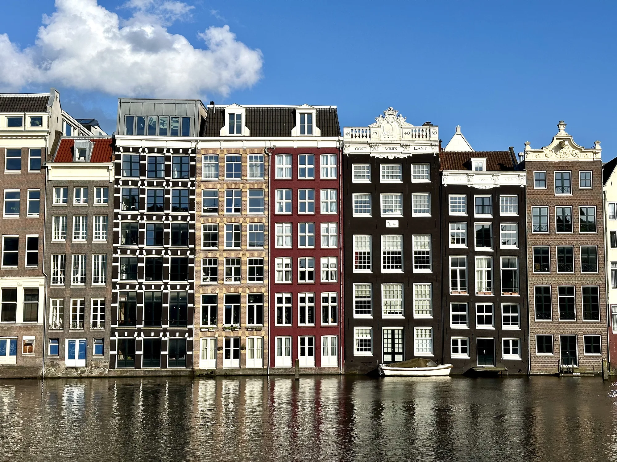 この写真は、オランダのアムステルダムの運河沿いに建ち並ぶ特徴的な家々を捉えています。運河の水面に家々のカラフルな外観が反射し、美しい景観を作り出しています。家々はそれぞれ異なる色合いを持ち、赤、茶色、黒など、多様な建築様式が混在しています。窓の数も多く、それぞれに白い枠が施されており、整然とした印象を与えています。一部の建物には装飾的な屋根や彫刻が施されており、歴史的な雰囲気を感じさせます。運河には小さなボートが係留されており、街の風景にアクセントを加えています。空は晴れており、白い雲が浮かんでいます。全体的に、アムステルダムの穏やかで美しい雰囲気が伝わってくる写真です。