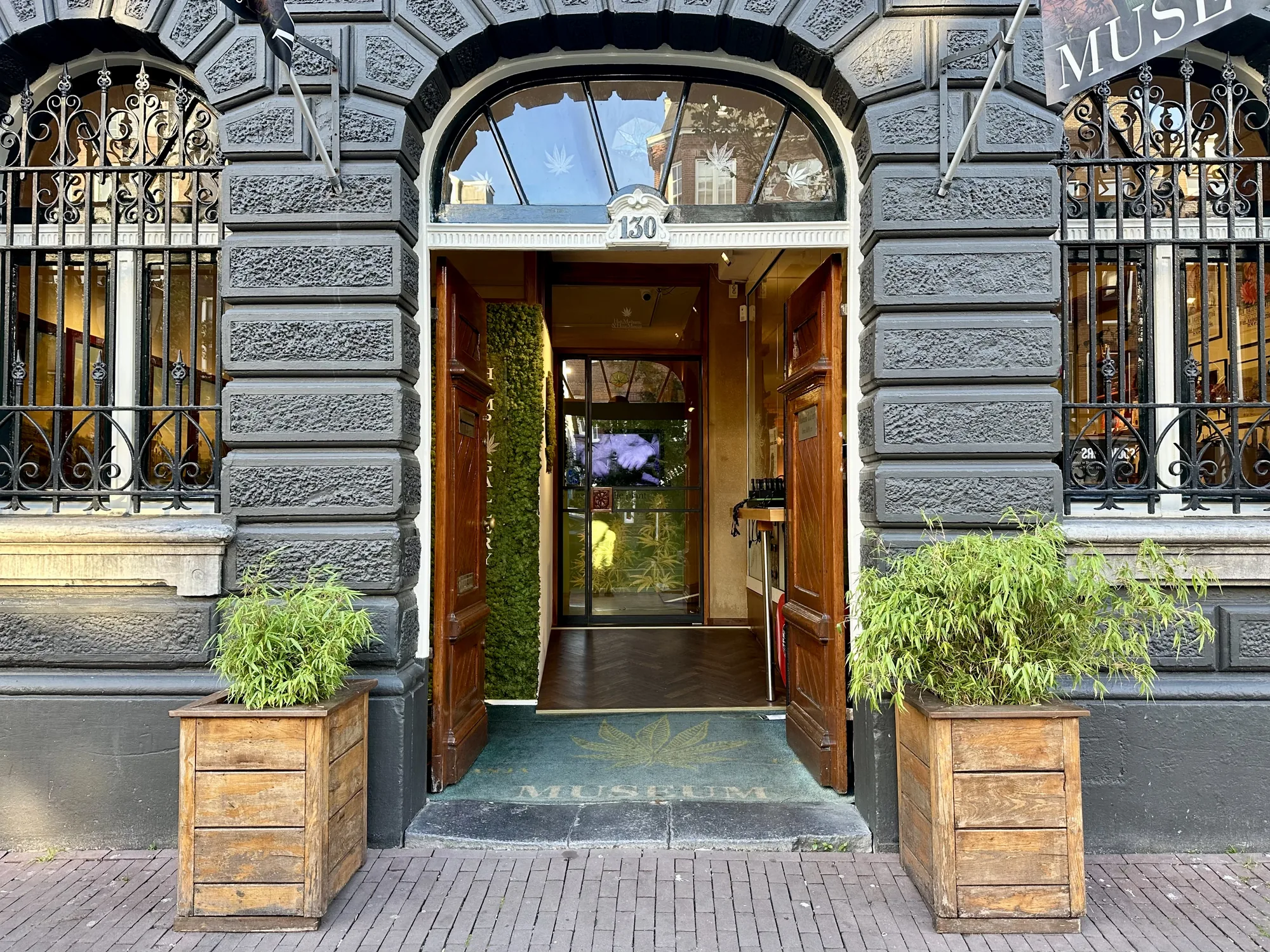アムステルダムにある大麻博物館の入口を捉えた写真です。重厚な石造りの建物の正面に、開け放たれた木製の扉が特徴的です。扉の上部には「130」と書かれた番号があり、アーチ型の窓には大麻の葉のマークが施されています。入口の床には博物館の名前が書かれたマットが敷かれ、建物の両脇には木製のプランターに植えられた植物が置かれています。窓には鉄格子が取り付けられ、内部には展示物と思われるポスターや写真が見えます。全体的に落ち着いた雰囲気で、歴史を感じさせる建物と大麻をテーマにした装飾が組み合わさっています。