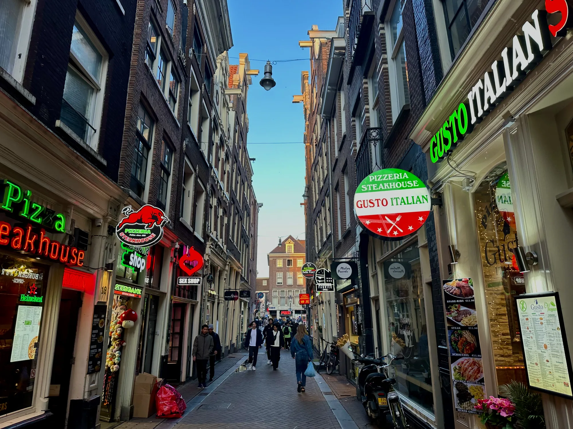 写真はオランダのアムステルダムで撮影された、狭い路地の風景を捉えています。路地の両側にはレンガ造りの建物が立ち並び、それぞれの建物には様々な店舗の看板が見られます。「Pizza Steakhouse」「Gusto Italian」といった飲食店や、「Sex Shop」といったお店のネオンサインが目を引きます。路地には数人の人が歩いており、その多くは観光客のように見えます。路地の奥には青空が広がっており、夕暮れ時の柔らかい光が路地全体を照らしています。建物は歴史を感じさせる古いレンガ造りのものが多く、趣のある雰囲気を醸し出しています。道は石畳で舗装されており、所々に自転車やバイクが停められています。全体的に活気があり、観光地らしい賑やかさを感じさせる写真です。