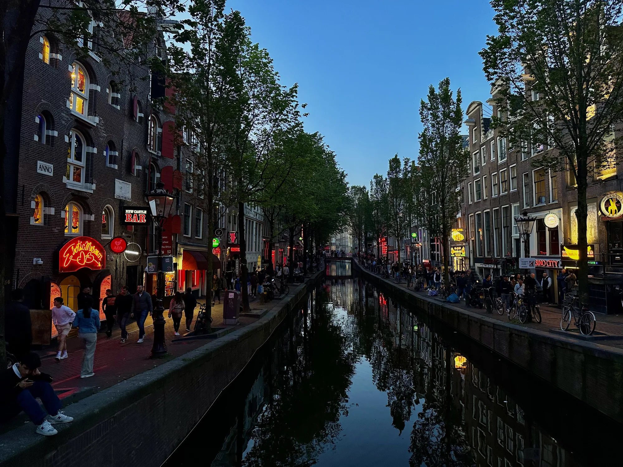 アムステルダムの運河沿いを捉えた写真です。時刻は夕暮れ時で、空は深い青色に染まっています。運河の両側には、レンガ造りの建物が立ち並び、ネオンサインが灯り始めています。通りには多くの人々が歩いており、賑わいを見せています。運河の水面には、建物やネオンの光が反射し、幻想的な雰囲気を醸し出しています。自転車が多数駐輪されている様子から、アムステルダムらしい風景が伝わってきます。写真全体からは、夜の始まりを迎え、これからさらに活気づいていく街の様子が感じられます。"Exotic Museum"や"New Castle Bar"といったネオンサインから、歓楽街の一面も垣間見えます。
