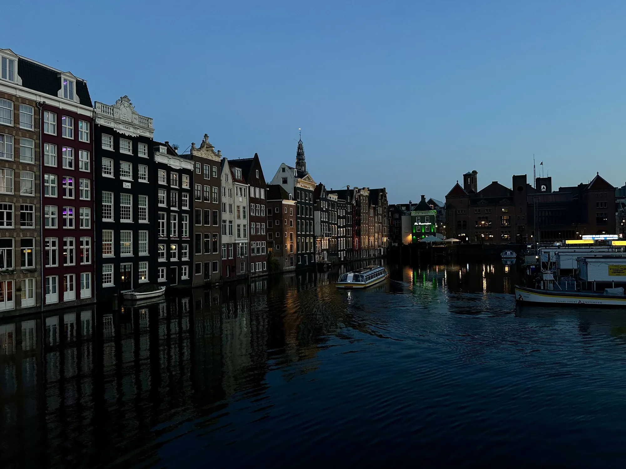 この写真はオランダのアムステルダムで撮影された、夕暮れ時の運河の風景です。空は深い青色に染まり、建物にはわずかに灯りがともり始めています。運河沿いには、伝統的な様式のカラフルな建物が立ち並び、その窓からは室内の光が漏れています。運河の水面は穏やかで、建物のシルエットや光が美しく反射しています。画面の中央には観光客を乗せた運河クルーズ船が航行しており、その光が水面に揺らめいています。右側には、停泊している観光船や建物が見えます。全体的に落ち着いた雰囲気で、アムステルダムの美しい夜景を捉えた一枚です。
