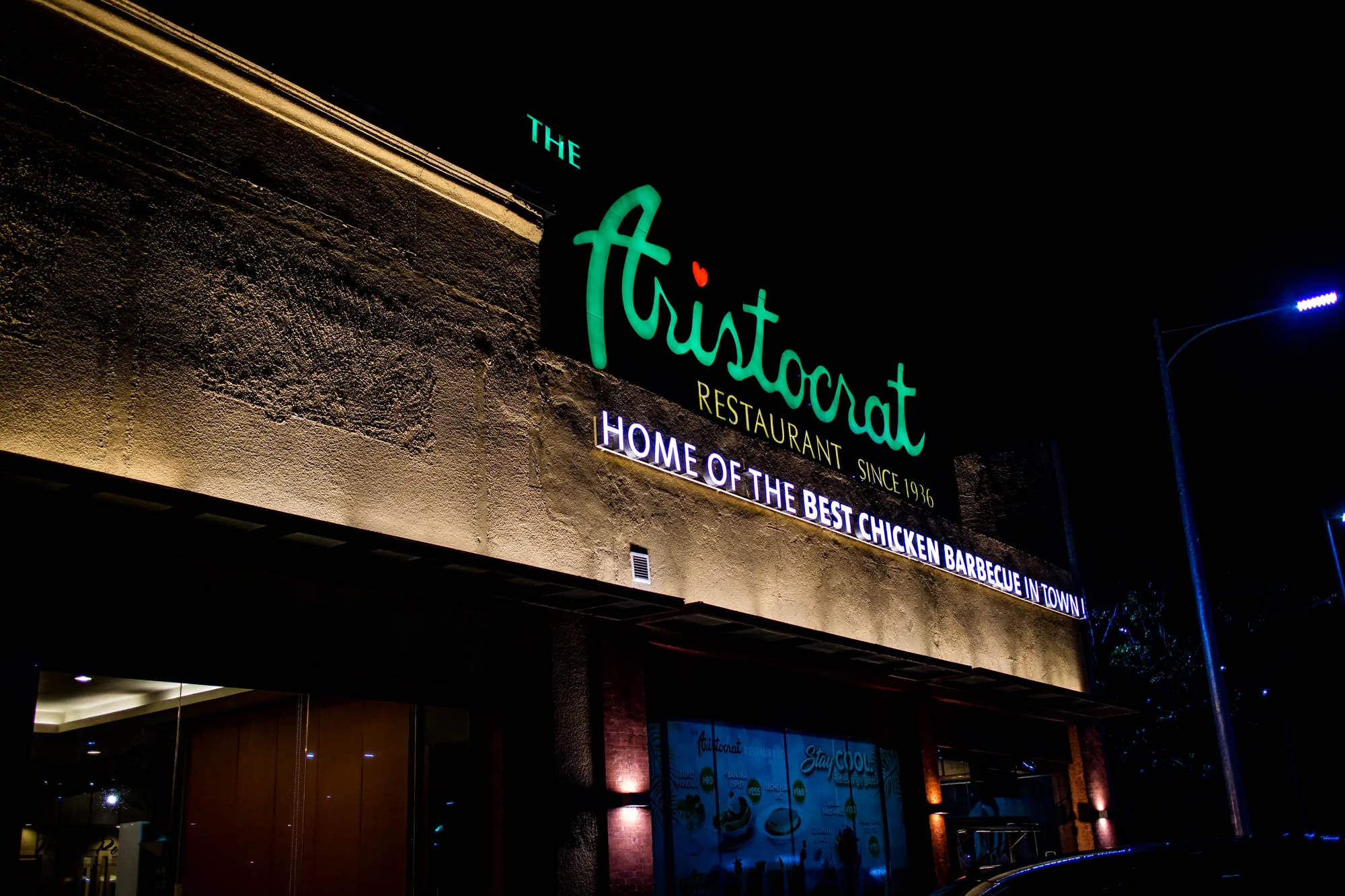 フィリピンのセブで撮影された夜景の写真です。写真には「The Aristocrat」というレストランの外観が写っています。緑色に光るネオンサインでレストランの名前が大きく表示されており、その下には「RESTAURANT SINCE 1936」と書かれた白いネオンサインがあります。さらにその下には、「HOME OF THE BEST CHICKEN BARBECUE IN TOWN!」という文字が同じく白いネオンで輝いています。  レストランの壁はベージュ色で、粗いテクスチャが特徴的です。壁の上部と庇の下には照明が設置されており、壁面を照らしています。店舗の入口付近にはメニューが掲示されており、写真からも「BUKO PANDAN P80」「BANANA SPLIT 255」「LECHE FLAN P160」といったメニューが読み取れます。  写真全体は夜間の撮影のため暗いですが、レストランのネオンサインが明るく輝き、周囲を照らしています。右奥には青色の街灯が写っており、夜の雰囲気を醸し出しています。