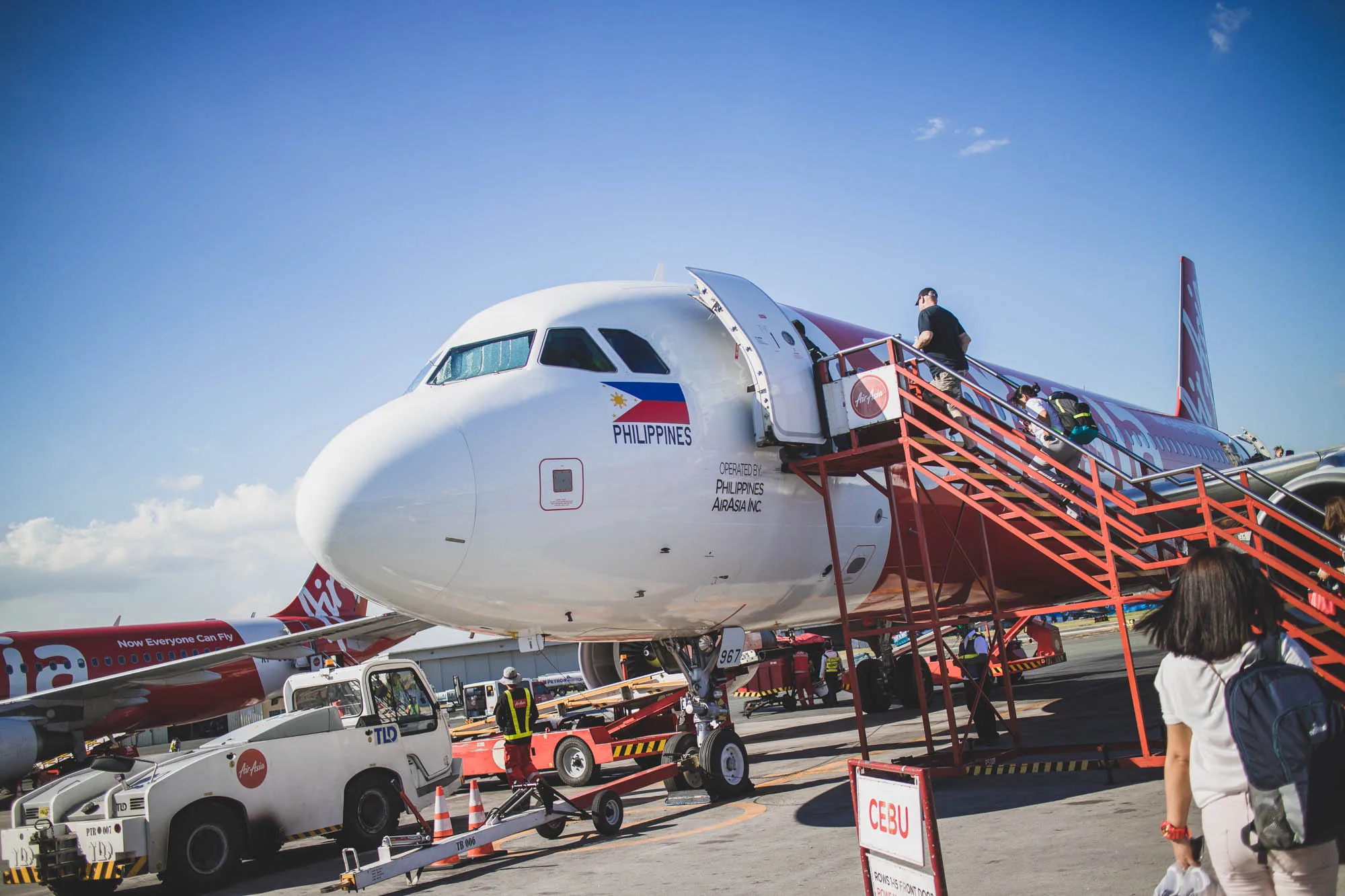 写真はフィリピンのセブ国際空港で撮影されたと思われる、エアアジアの飛行機の様子を捉えています。空は青く晴れており、機体は白と赤を基調としたエアアジアのデザインです。機体にはフィリピンの国旗が描かれており、「PHILIPPINES」の文字と、運営会社である「PHILIPPINES AIRASIA INC.」の文字が見えます。  赤い階段が機体に取り付けられており、数人の乗客が搭乗している様子が写っています。階段の近くには、空港作業員と思われる人物が立っており、機体の下に牽引車のような車両が確認できます。  写真の手前には、搭乗を待つ乗客の後ろ姿が見えます。看板には「CEBU」と書かれており、場所がセブであることを示唆しています。全体的に、旅行や空港の雰囲気を伝える写真です。