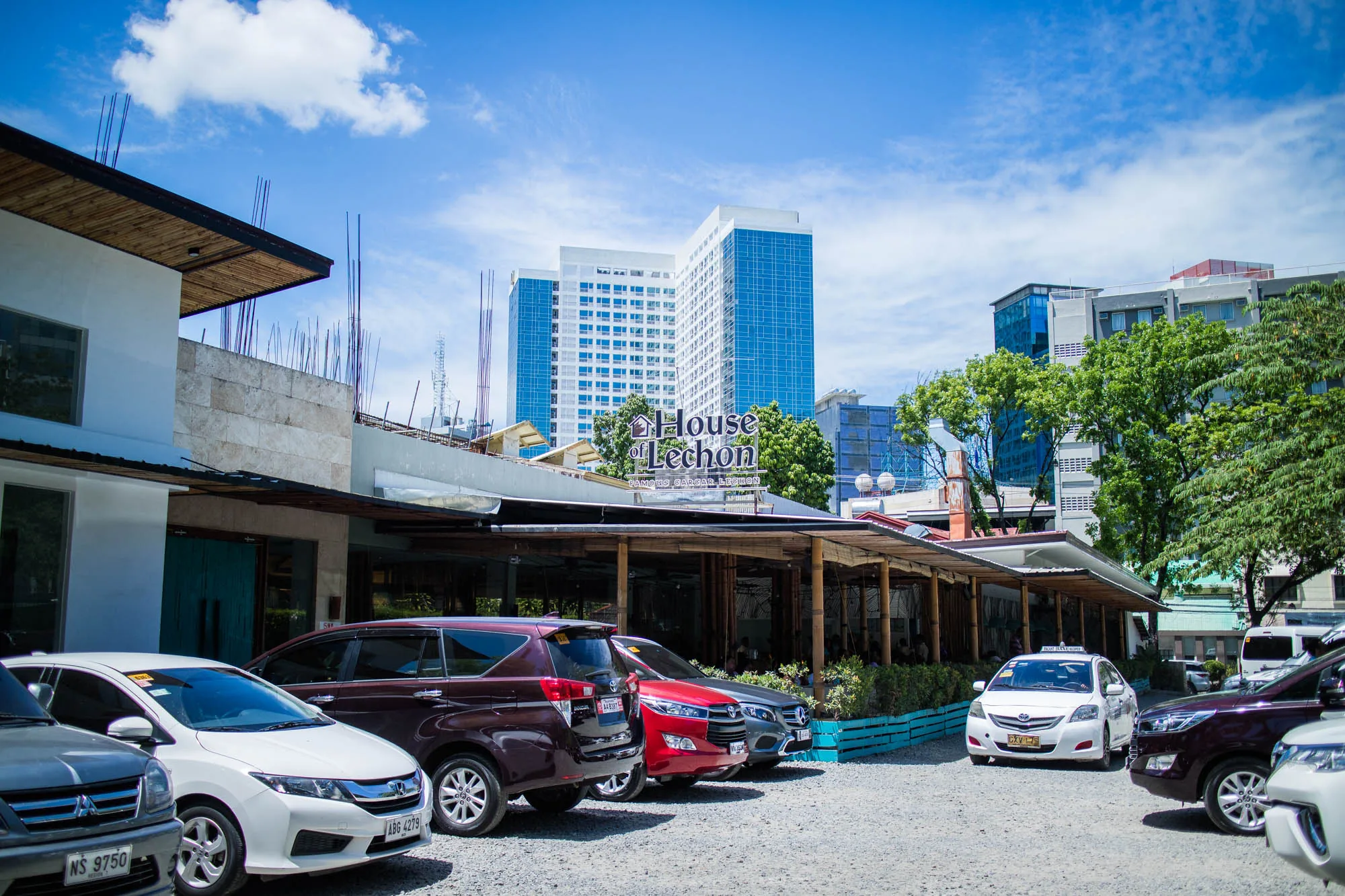 フィリピンのセブで撮影された写真です。手前には砂利の駐車場があり、様々な車種の車が並んでいます。駐車場奥には「House of Lechon」と書かれた看板を掲げたレストランがあり、多くの人で賑わっているようです。レストランの建物は竹でできた柱と屋根で覆われており、開放的な雰囲気です。背景には高層ビルが建ち並び、青い空には白い雲が浮かんでいます。日差しが強く、明るい写真です。ローカルの人気レストランの賑わいを伝える写真と言えるでしょう。