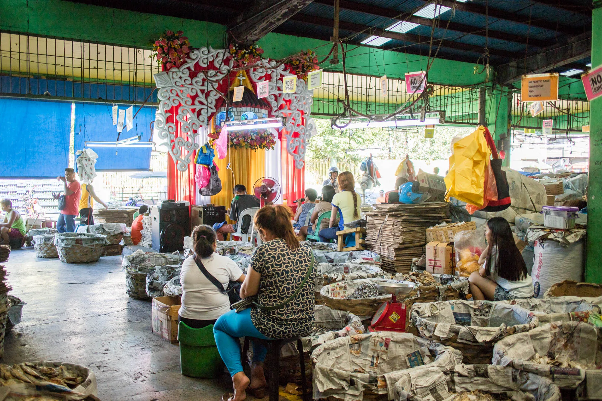 写真はフィリピンのセブで撮影された市場の様子を捉えています。屋内と思われる場所で、天井は緑色で錆びた金属製の骨組みが見えています。手前には、新聞紙が敷き詰められた籠が多数並んでおり、中には干物のようなものが入れられています。籠の周りには、女性たちが腰掛けており、商品を整理したり、計量器を使って量ったりしているようです。  写真中央奥には、装飾された祭壇のようなものがあり、花やリボン、飾りなどが施されています。その前には人々が集まっており、何かのイベントが行われているのかもしれません。奥の壁は青いカーテンで覆われていますが、所々隙間から外の光が差し込んでいます。  写真全体はやや暗く、市場の活気と雑多な雰囲気が伝わってきます。天井から吊り下げられた看板や、商品が積み上げられた様子から、生活感あふれる場所であることがわかります。